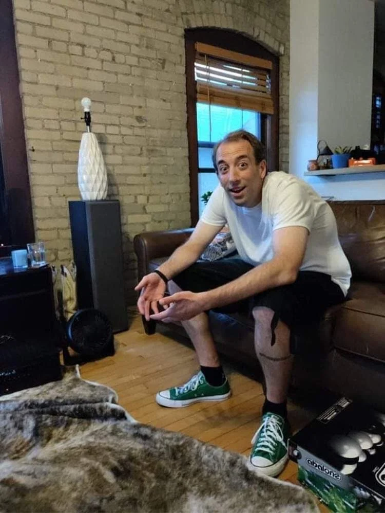 A man sitting on a brown leather couch in a living room with a brick wall, window, and wooden floors, wearing a white t-shirt, black shorts, and green sneakers, gesturing with his hands and looking surprised or excited.