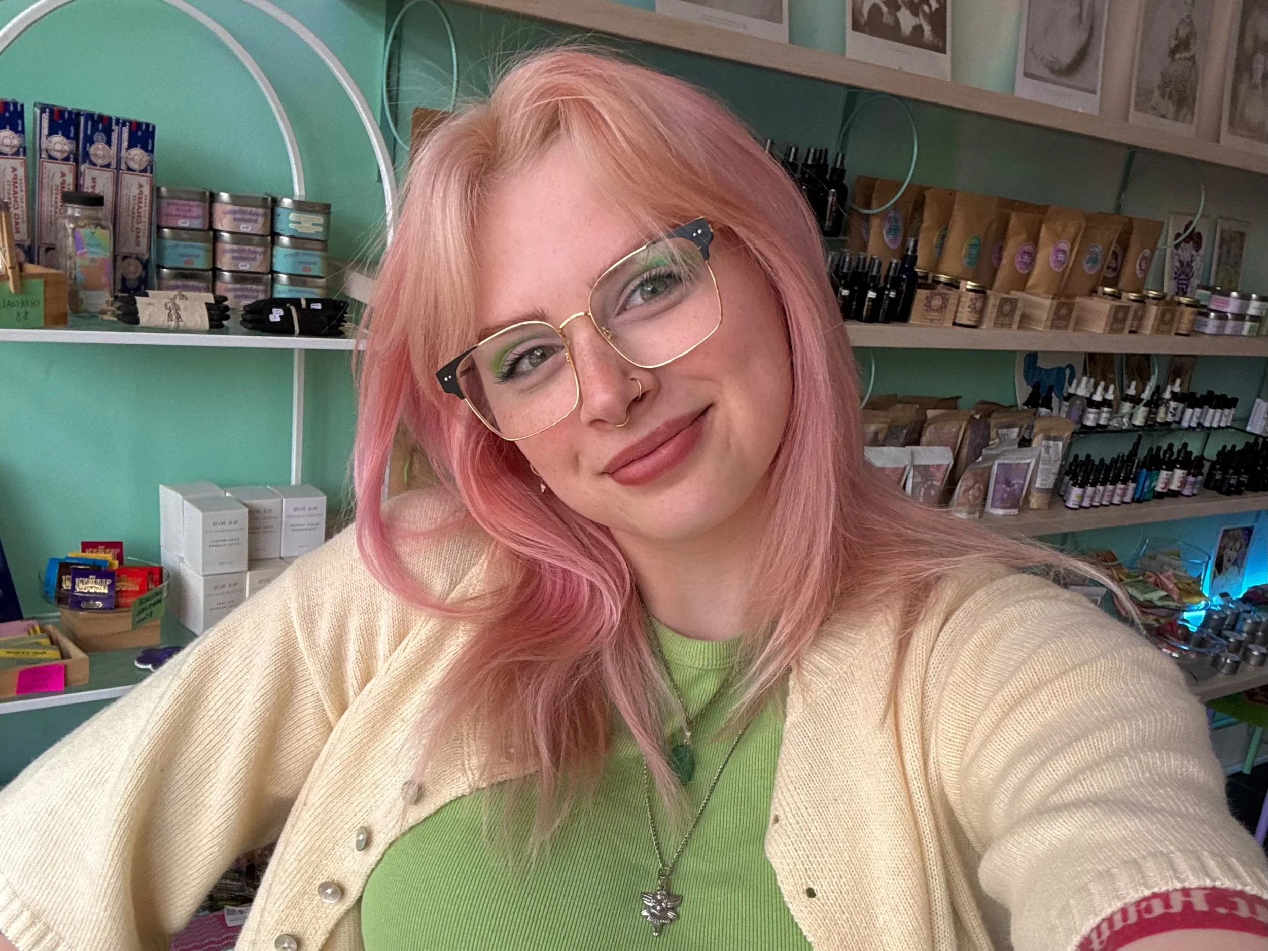 A young woman with pink hair, wearing glasses, a beige cardigan, and a green shirt, smiling inside a store with shelves of products.
