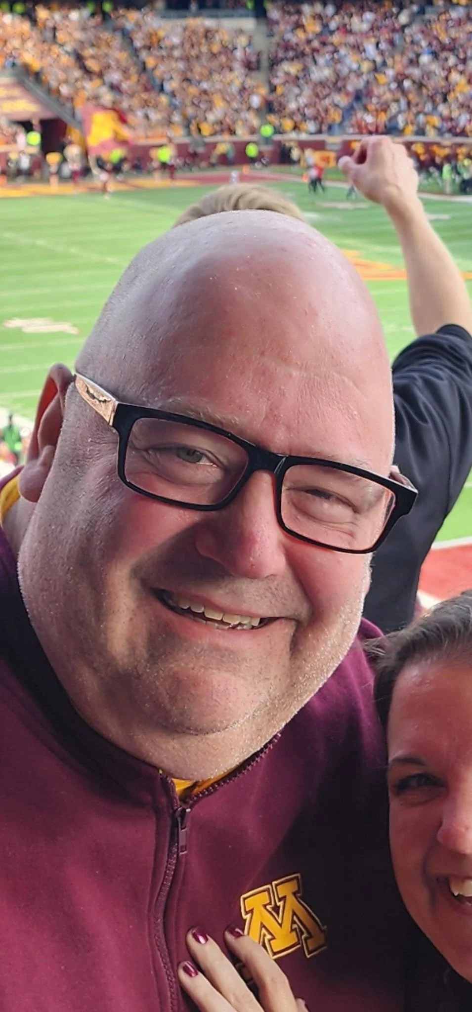 A close-up of a smiling man with glasses and a bald head at a football stadium, with a football field and crowd in the background.