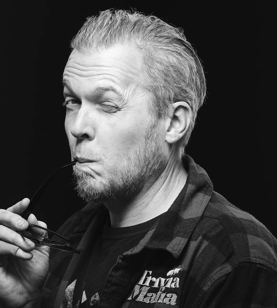 Black and white portrait of a man with short, slicked-back hair and a goatee, holding glasses and making a playful, winking facial expression.