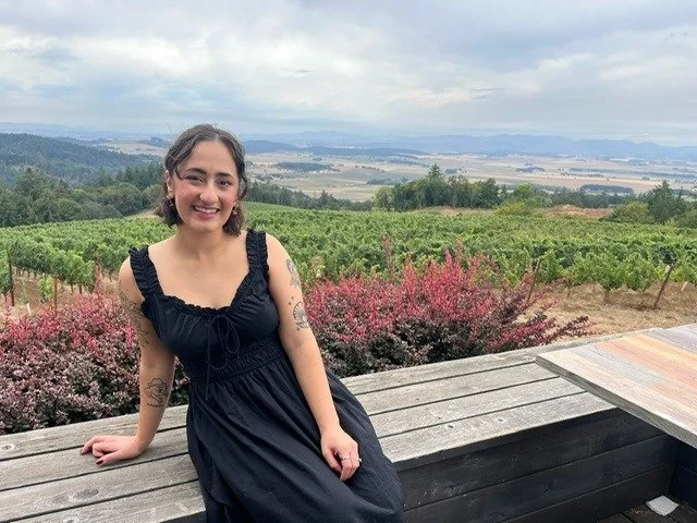 Young woman with short brunette hair, wearing a black dress, and sitting on a wood bench in front of a scenic background