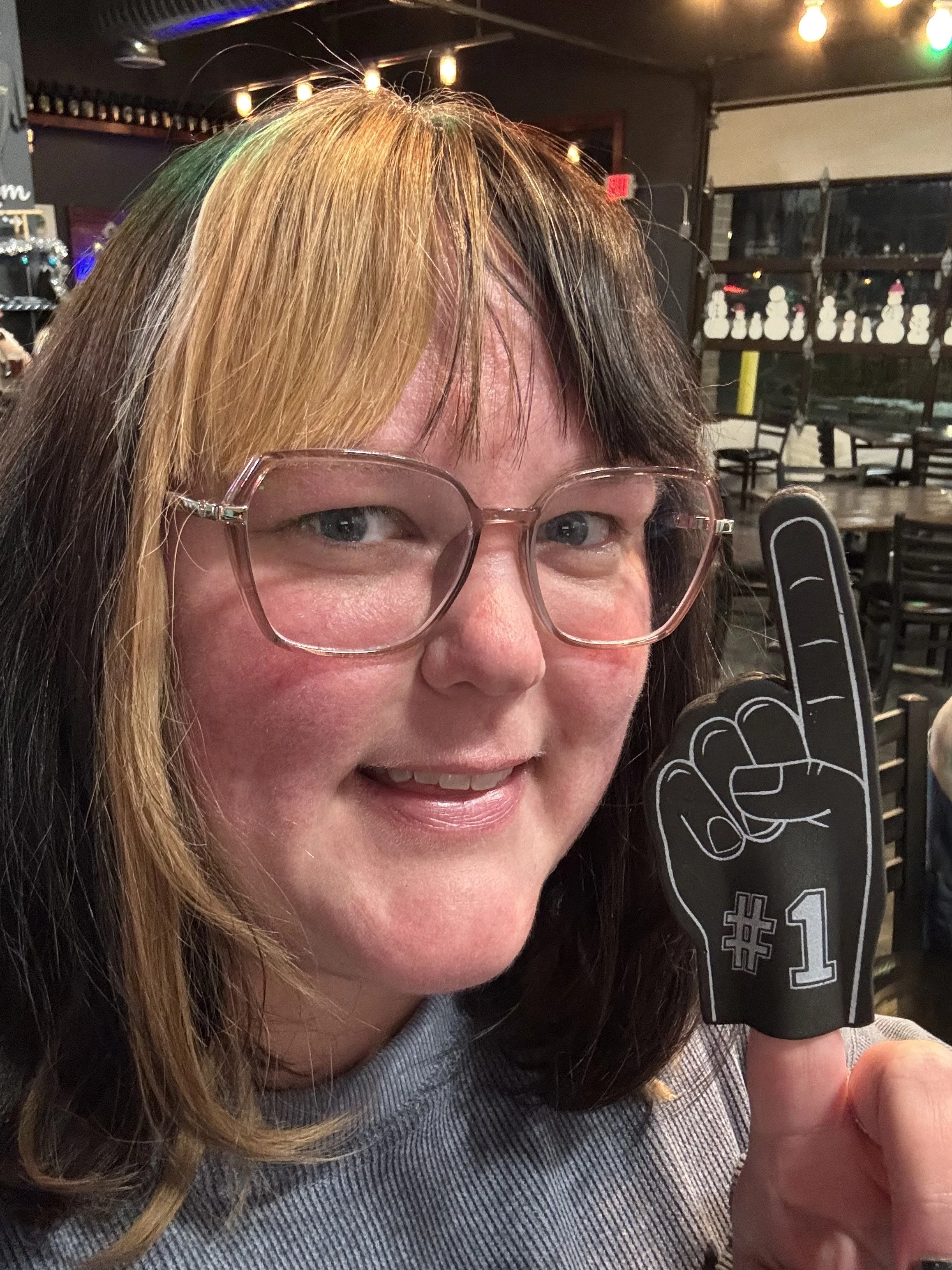 A woman with glasses and shoulder-length hair that is blonde on one side and dark on the other, smiling while holding up a foam hand foam finger with the #1 symbol.