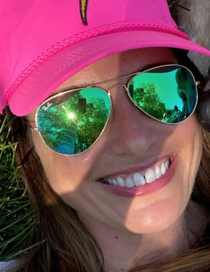 A woman smiling outdoors wearing a pink baseball cap and reflective aviator sunglasses.