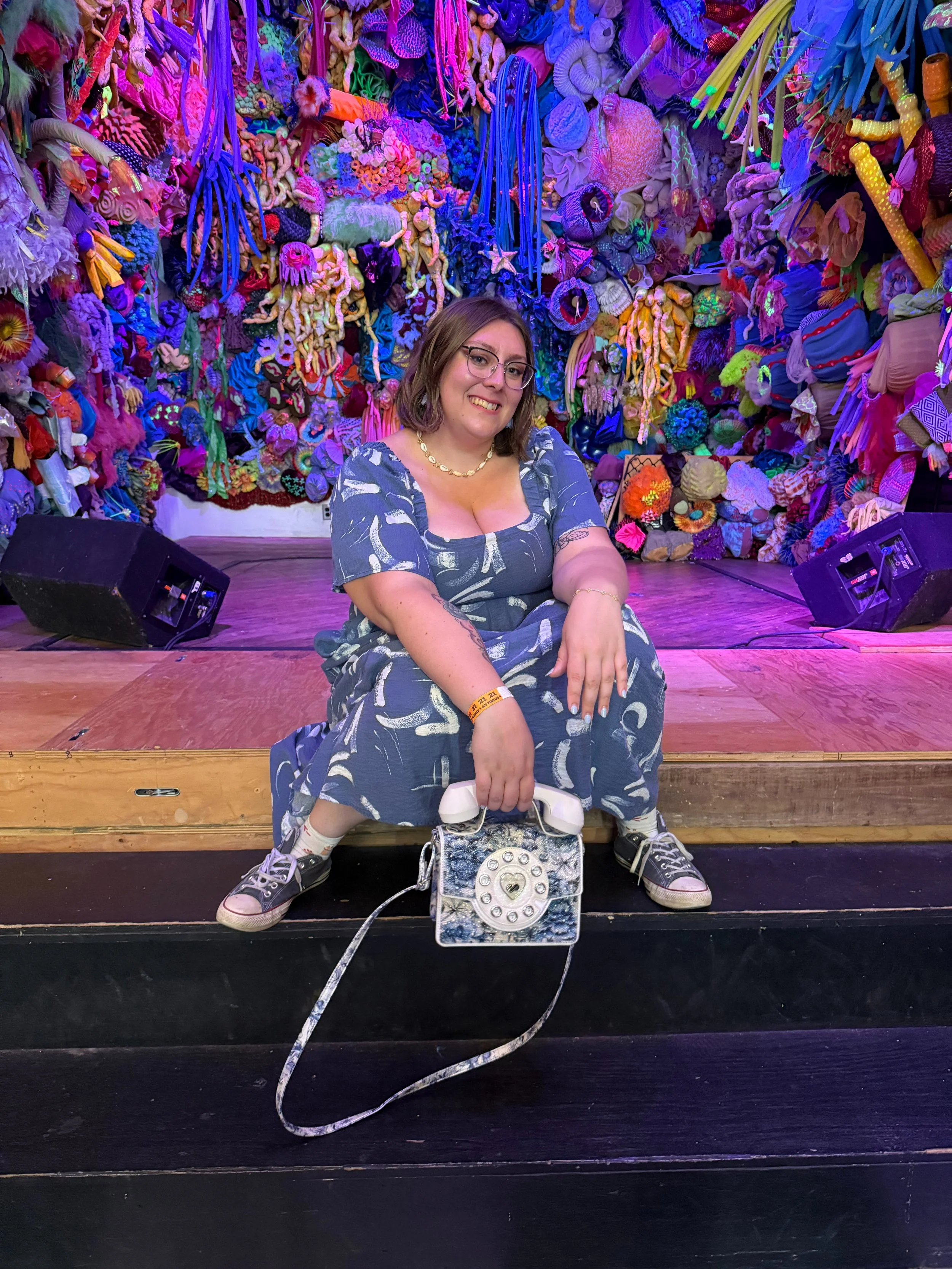 A woman wearing a blue dress with white patterns, glasses, and sneakers, sitting on a black stage step, holding a vintage-style purse, in front of a colorful background made of various textiles and fabric art pieces in bright colors.