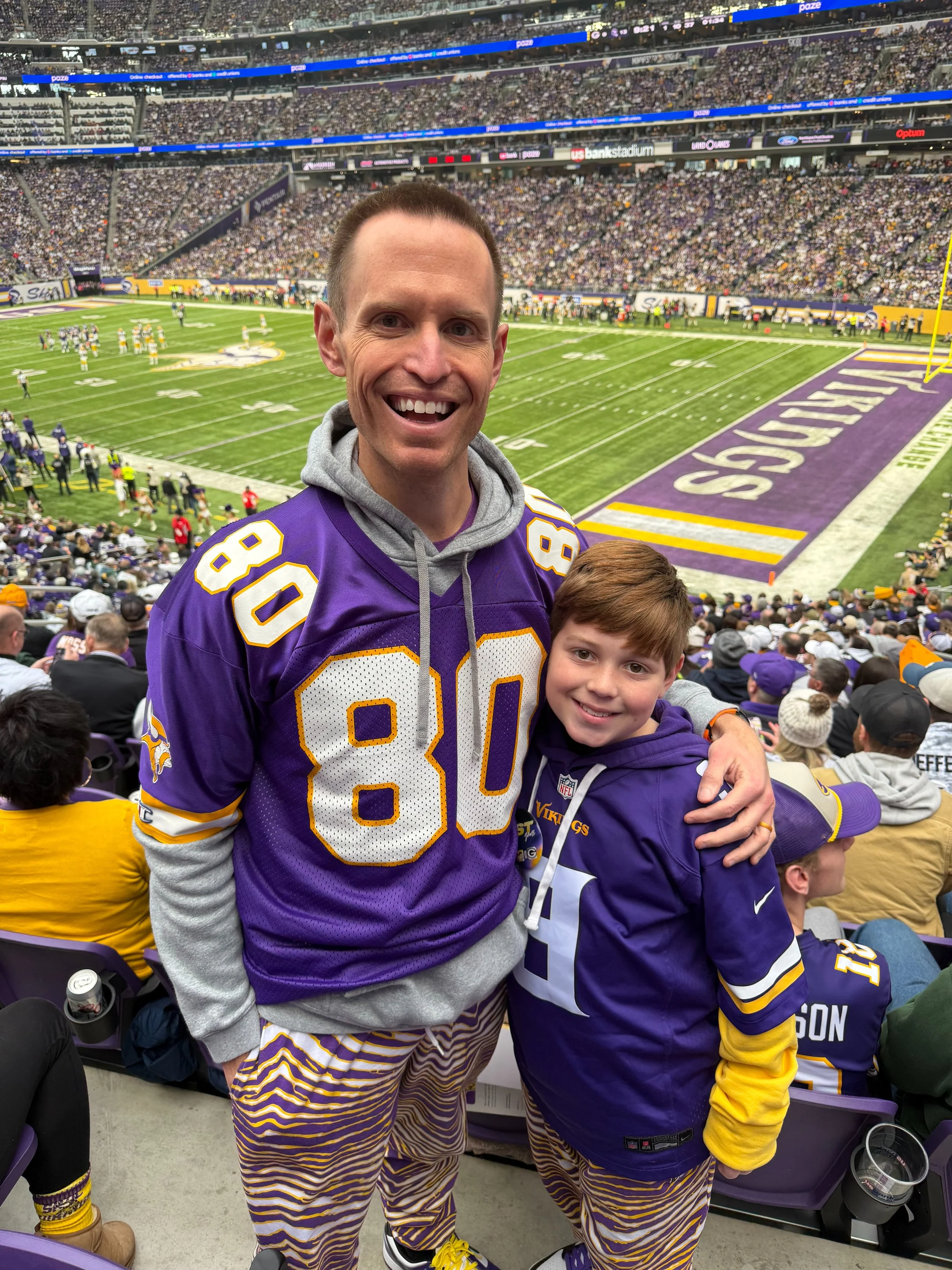 Two fans at a Vikings football game, smiling, wearing purple and yellow team jerseys and matching striped pants, sitting in the stadium with the field in the background.