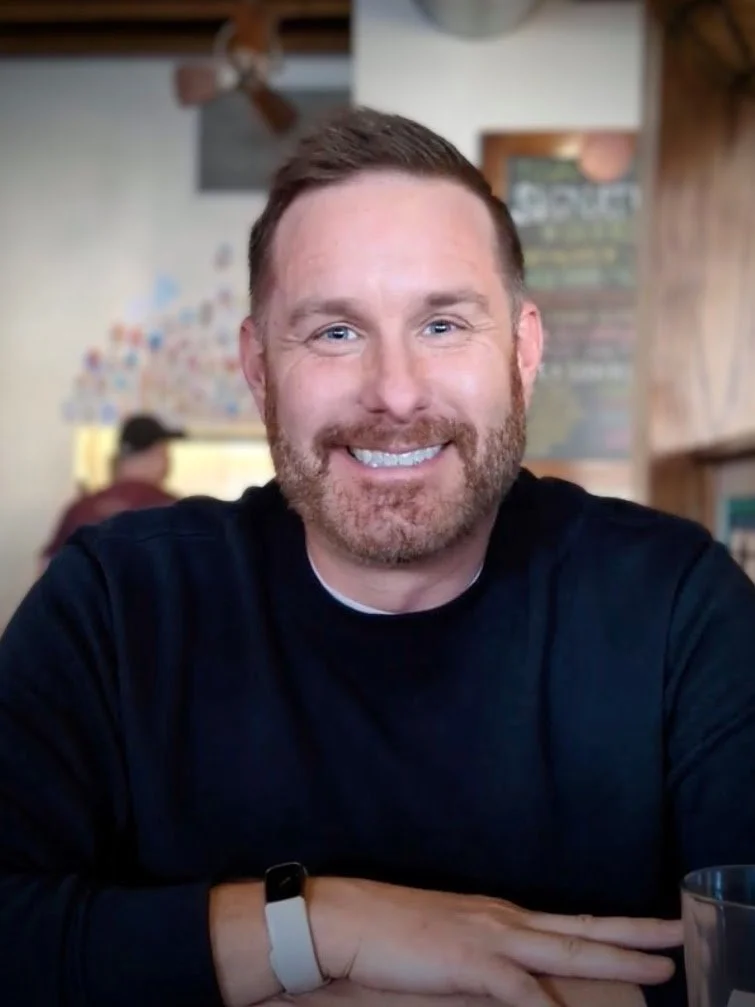 A smiling man with a beard and short hair, wearing a black shirt, sitting at a table in a restaurant or cafe.