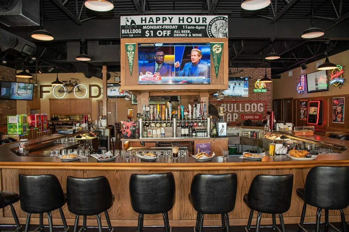 A bar inside a restaurant with a wooden counter, several black chairs, multiple beer taps, and TVs hanging from the ceiling showing sporting events and news, with signs for happy hour and food and drink options.