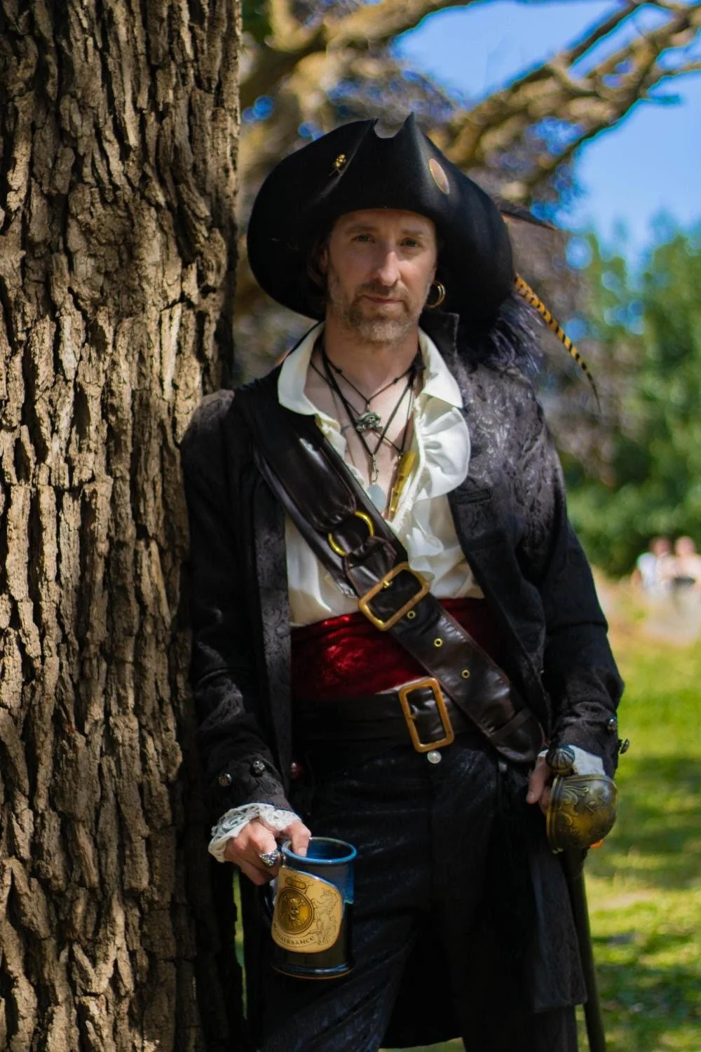Man wearing an authentic pirate outfit, standing against the trunk of a tree and holding a mug of beer