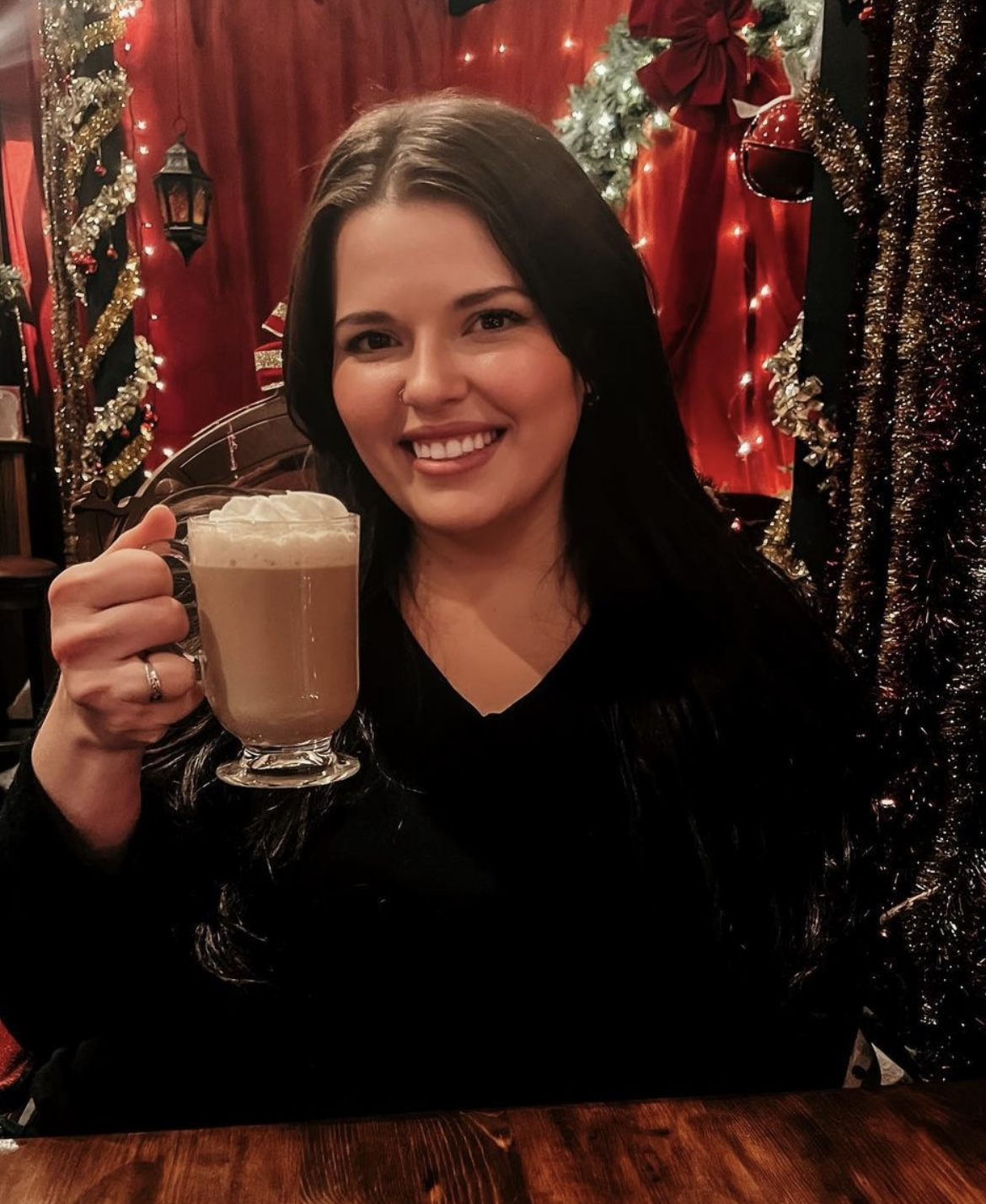 A woman with long dark hair and smiling face holding a cup of coffee or hot chocolate in a decorated festive setting with Christmas lights and ornaments in the background.