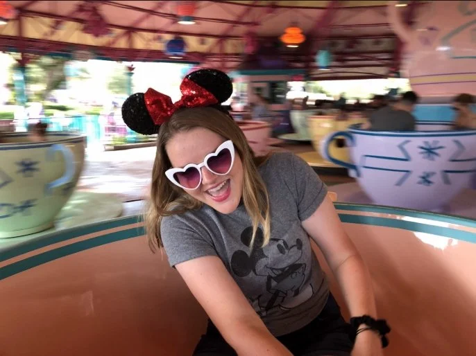 A woman sitting in a teacup ride at an amusement park, wearing heart-shaped sunglasses and Minnie Mouse headband with red bow, smiling and laughing.