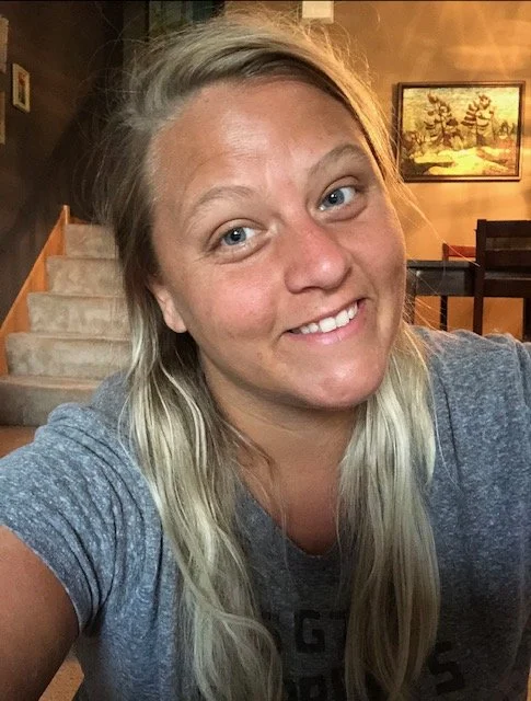 A young woman with long, blonde hair smiling at the camera in a cozy home setting with a staircase and framed pictures on the wall behind her.