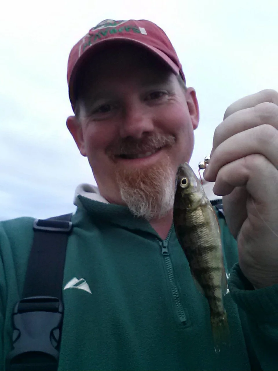 A man wearing a red cap and green jacket holding a small fish, smiling at the camera outdoors.