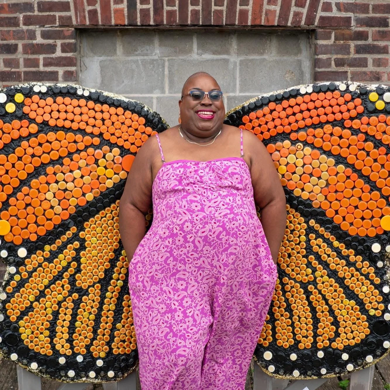 A smiling person with glasses and a pink floral dress standing in front of a butterfly-shaped art piece made of orange and black bottle caps, with a brick wall in the background.