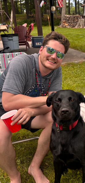 A man smiling while sitting on the grass with a black dog, holding a red plastic cup in his hand. The background shows outdoor patio furniture, flags, and trees.