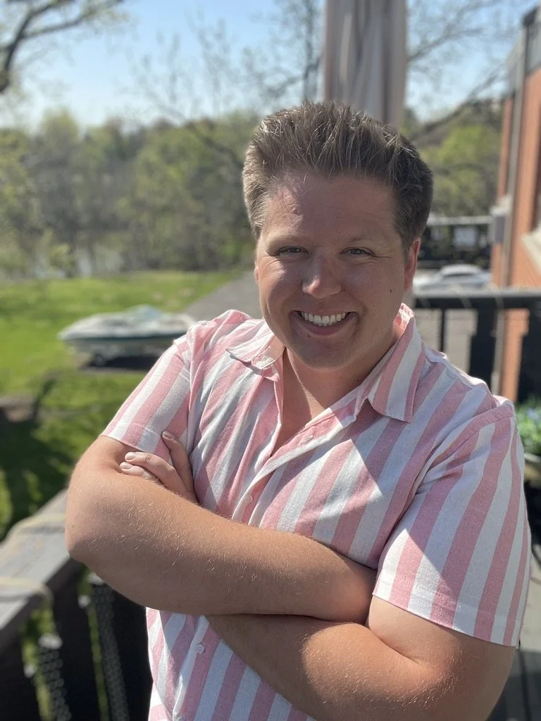 Guy standing on a porch and smiling with his arms folded in front of his chest