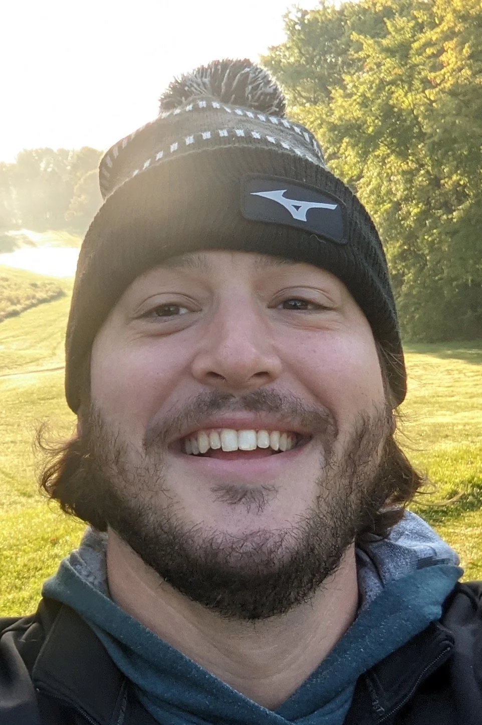 A smiling man taking a selfie outdoors in a grassy area with trees and a small body of water in the background, wearing a black beanie with a logo and a hoodie.