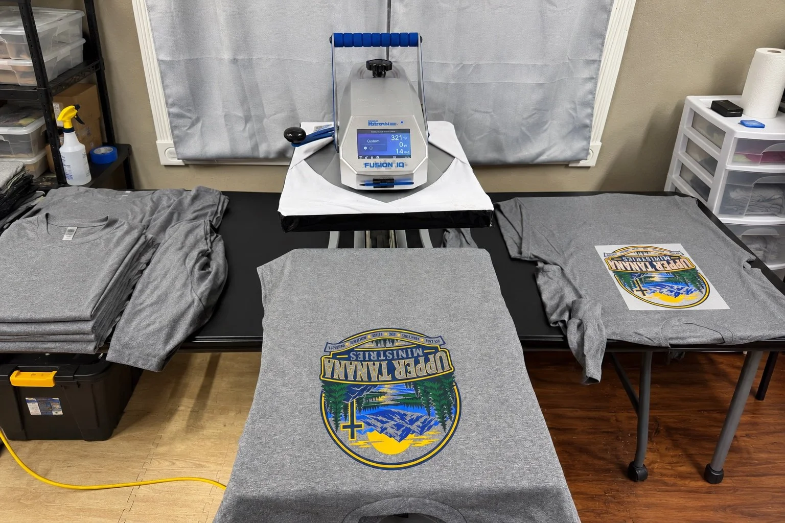 Pressing a gray t-shirt with a mountain and lake logo on a heat press machine in a room. Several other gray shirts are folded and stacked to the left, and one is partially pressed with the logo on it. To the right, there is a plastic drawer unit and a spray bottle on a shelf.