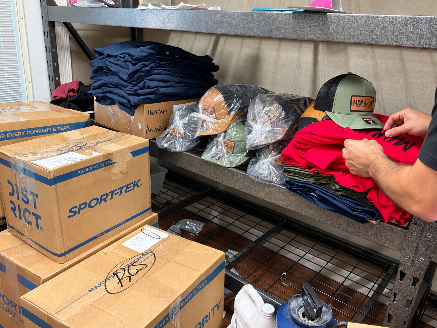 Shelf with organized clothing items including boxed sportswear, hats in plastic bags, and red, blue, and black folded shirts or pants, with a person holding a red shirt and a green cap with a patch that reads 'Men Arise'.