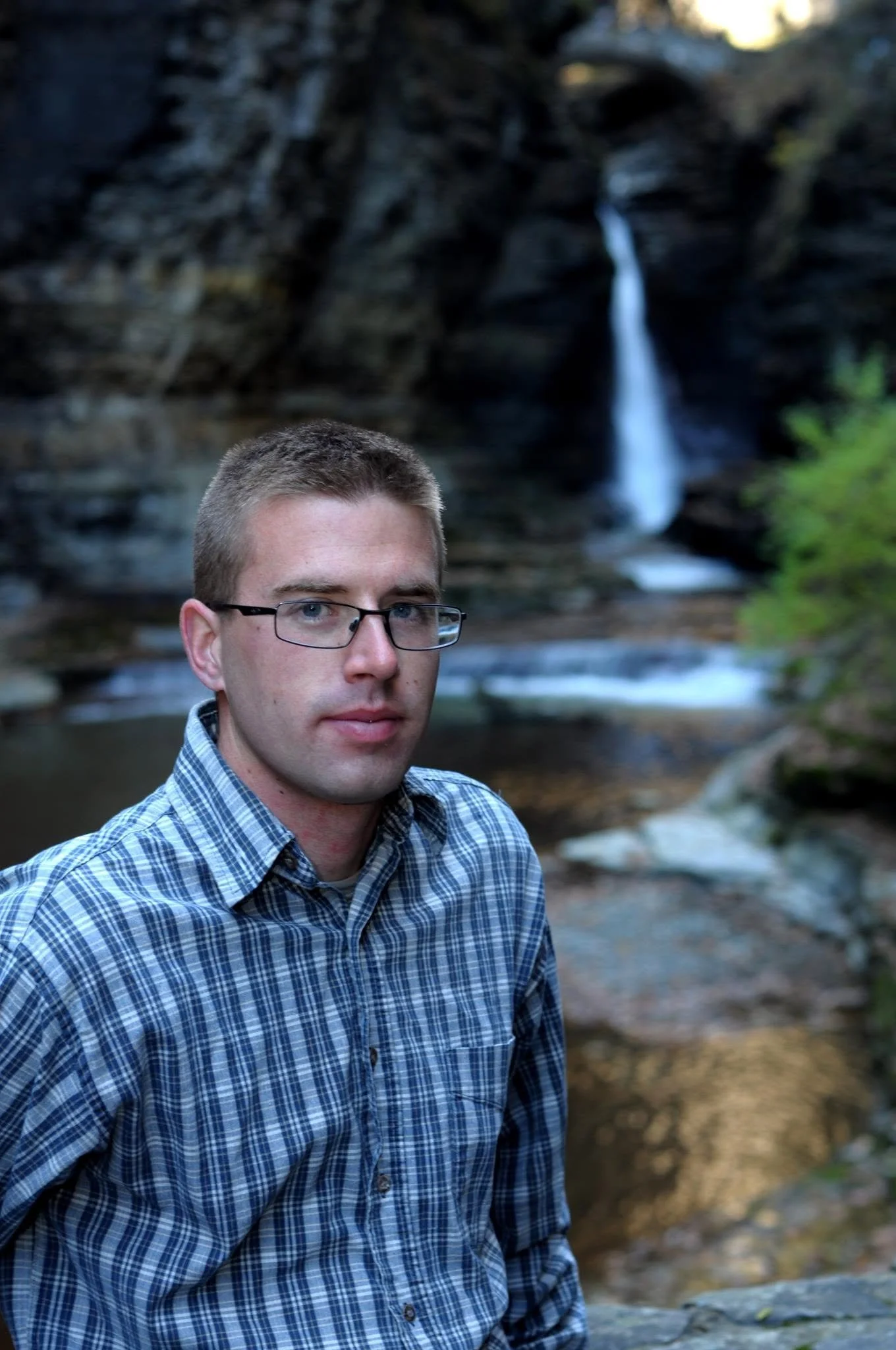 A man with short hair, glasses, and a blue plaid shirt standing outdoors in front of a rocky waterfall and greenery.