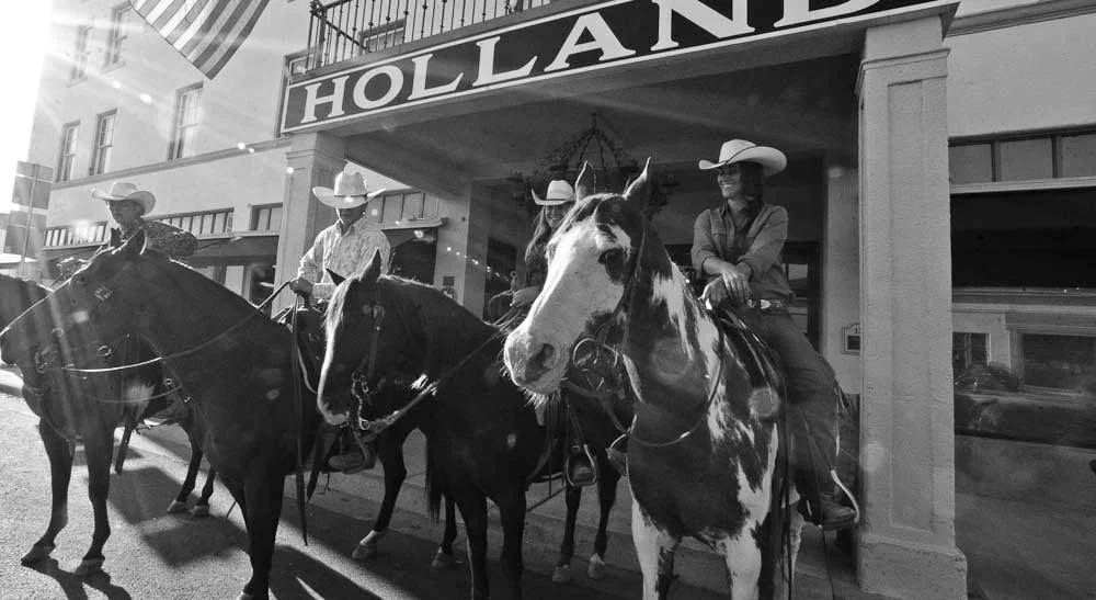 A black and white historical photo of four riders on their horses posing in front of Holland Hotel in Alpine, Texas.