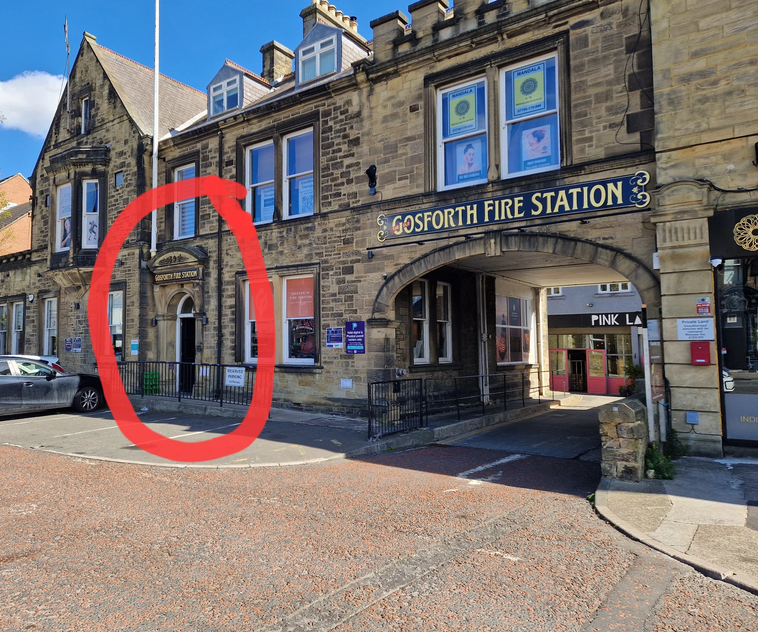 A stone building with a sign reading 'GOSFORTH FIRE STATION' and an arched driveway entrance. Several cars are parked in front. The sky is blue with some clouds.