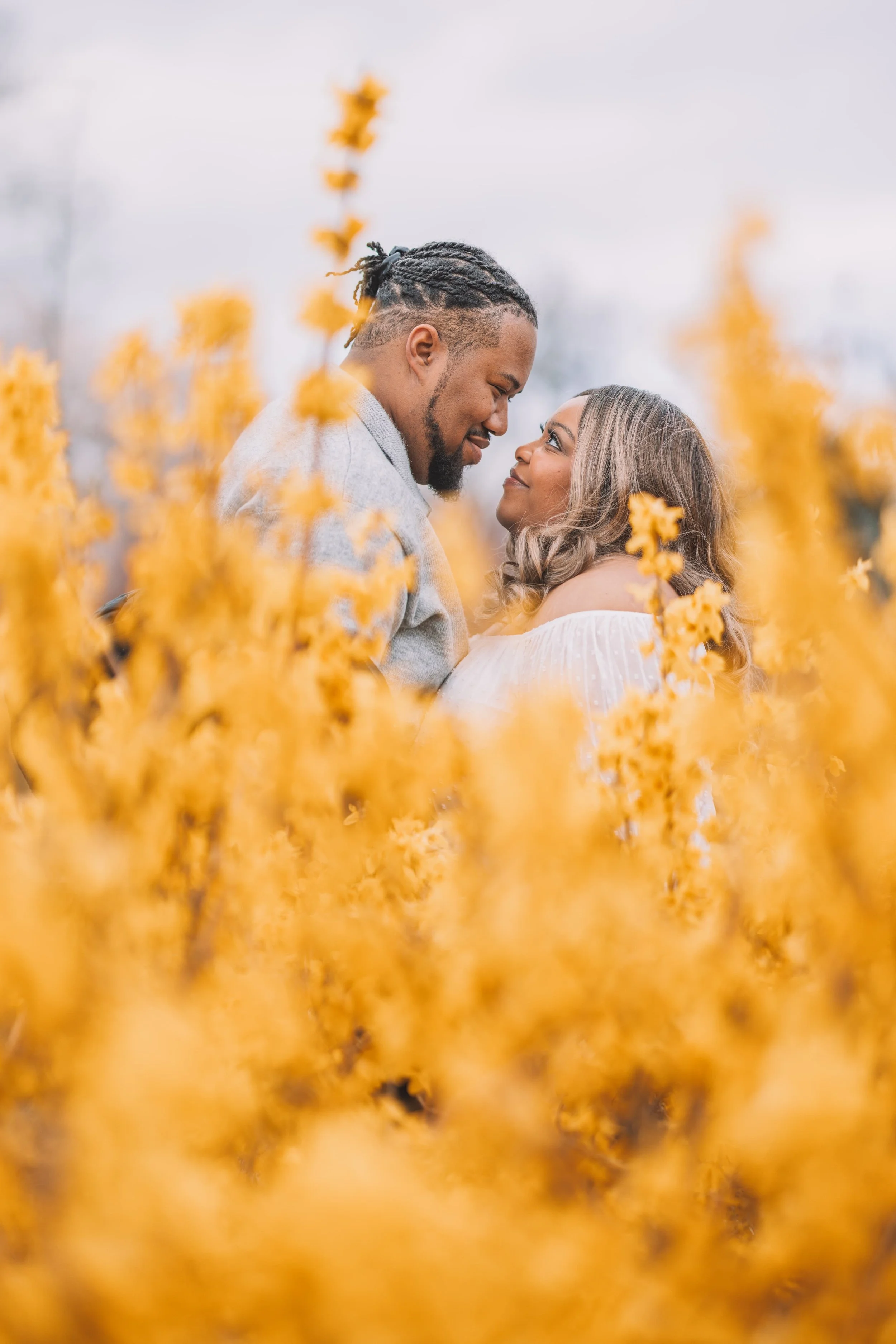 A couple standing close together in a field of yellow flowers, gazing into each other's eyes with a blurred background.