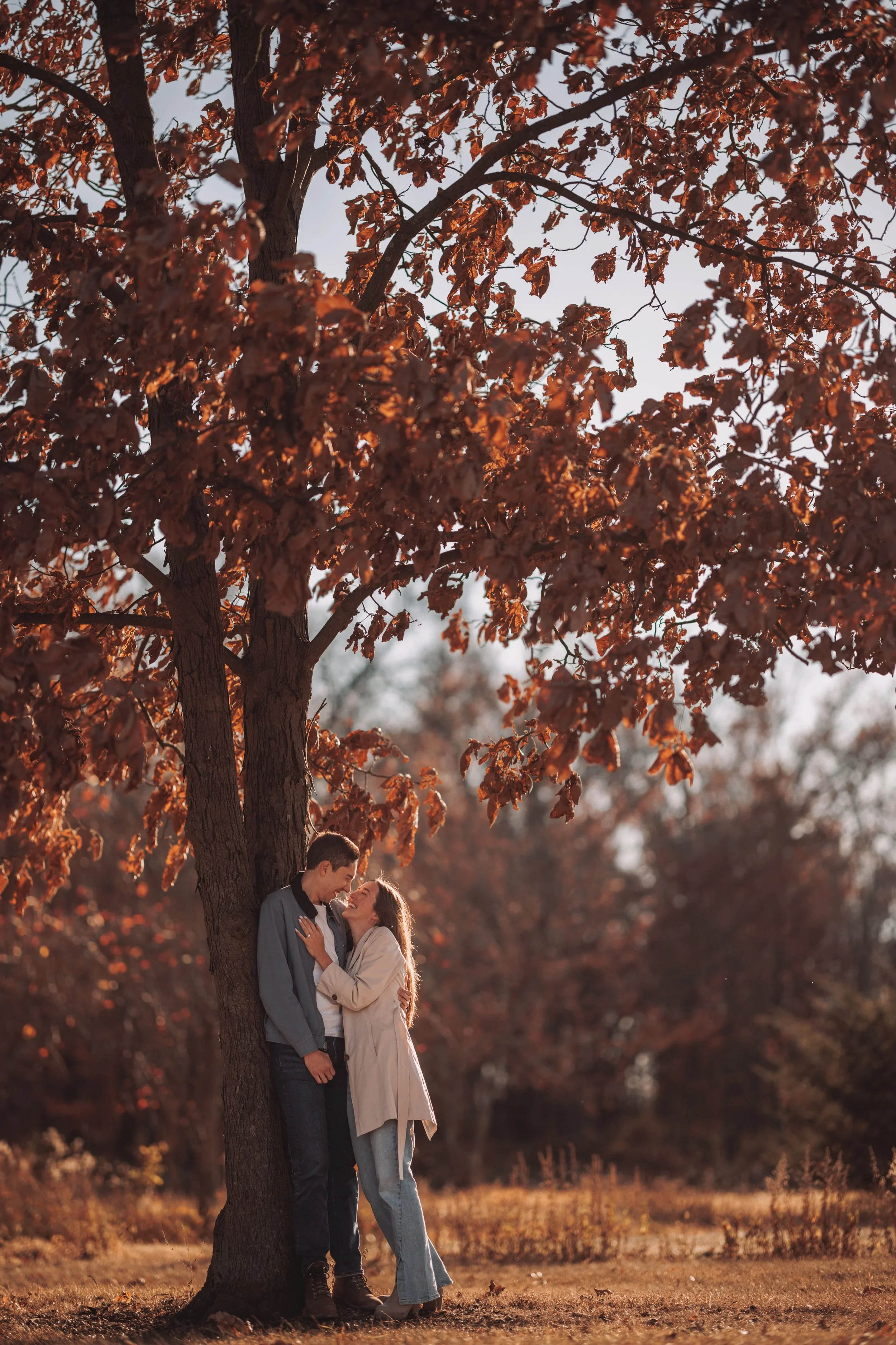 A couple standing close together under a large tree with brown leaves during fall, sharing an intimate moment in an outdoor setting.