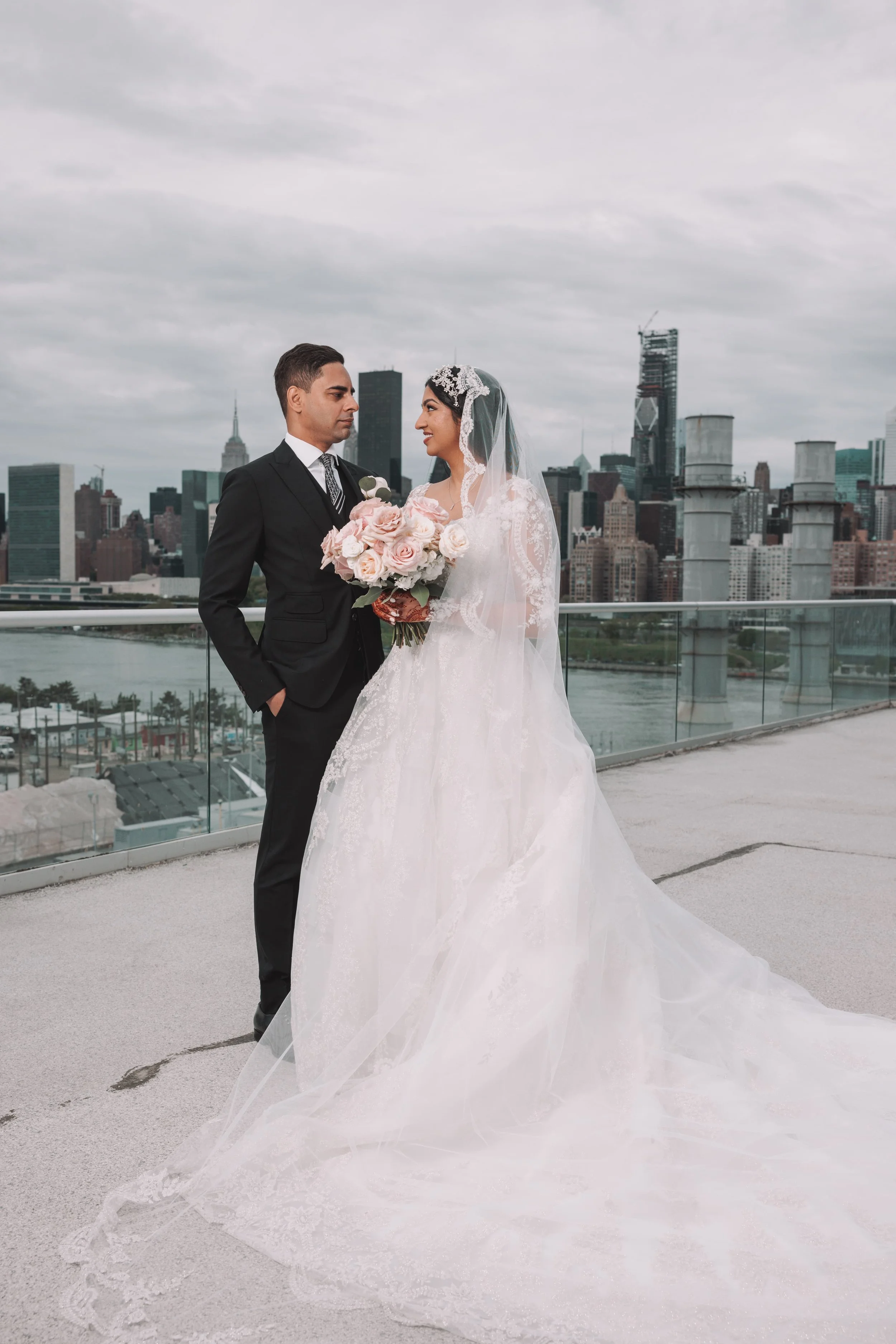A bride and groom stand on a rooftop with the New York City skyline in the background. The bride holds a bouquet of pink and white roses, wearing a white lace wedding gown and veil. The groom wears a black suit with a tie, looking at the bride.