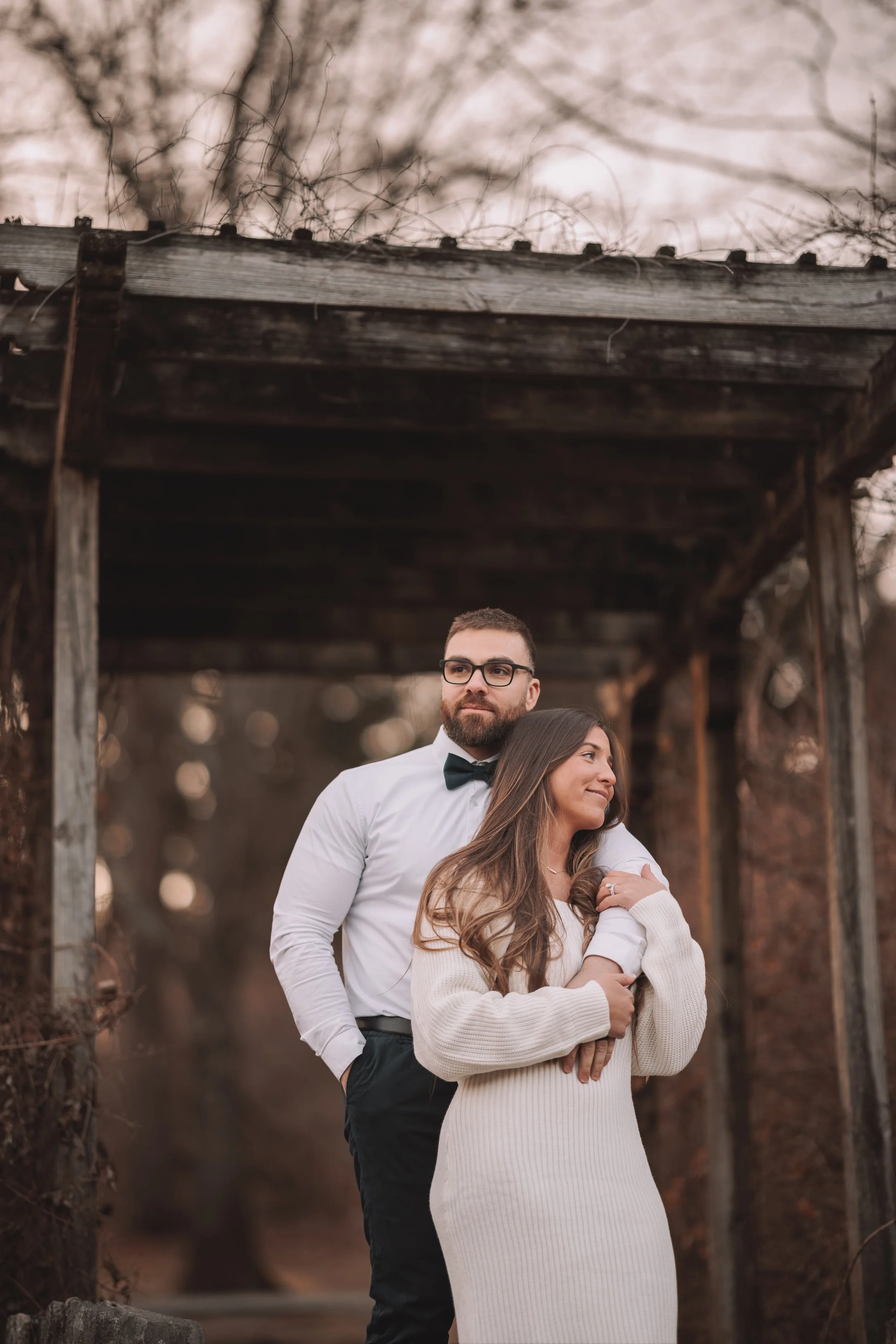A man in a white shirt, black pants, and bow tie stands behind a woman in a cream-colored knit dress, both are posing outdoors under a wooden structure during sunset.
