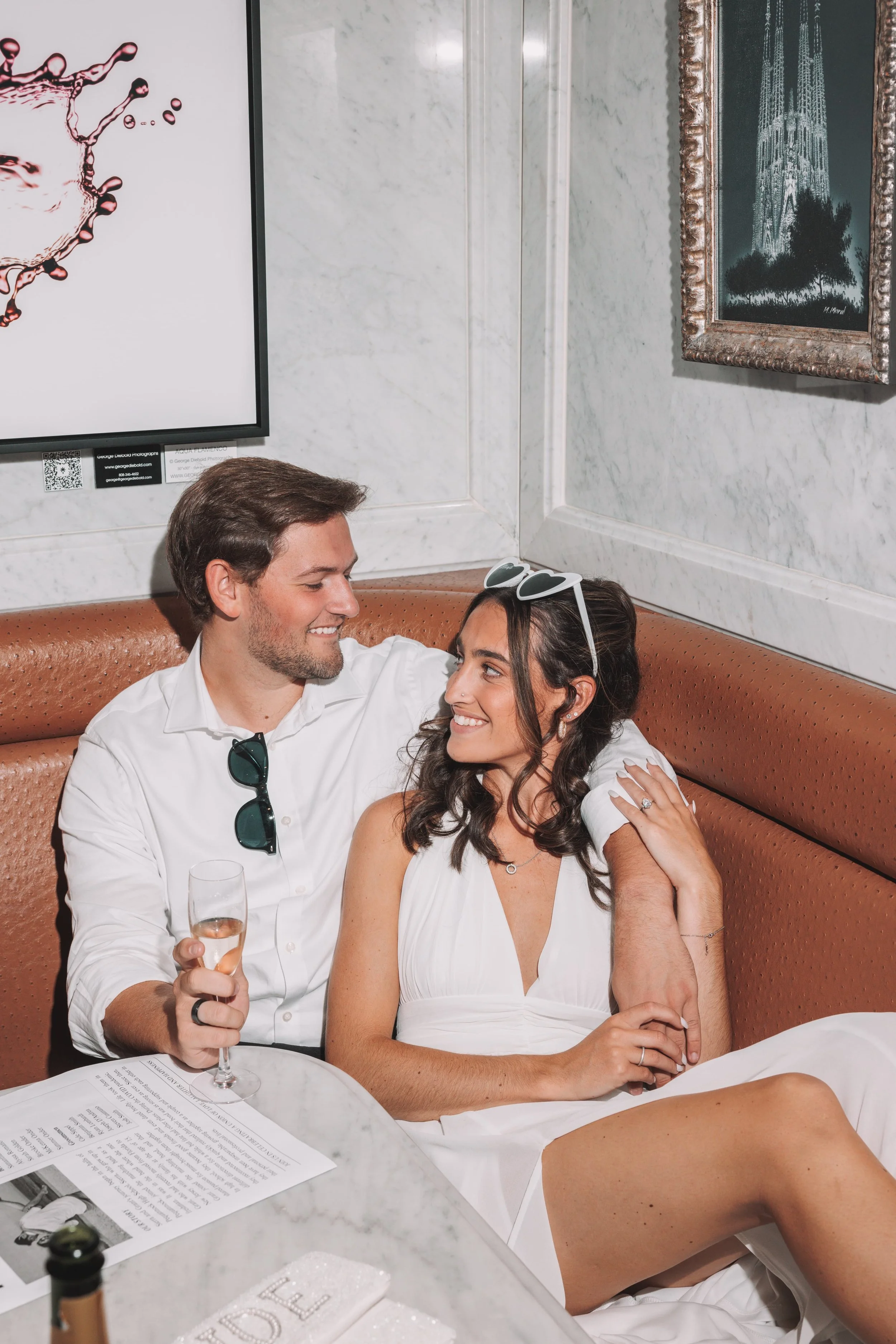 A smiling couple sitting close together in a restaurant, with the man holding a glass of champagne, and the woman wearing sunglasses on her head and showing a wedding ring.