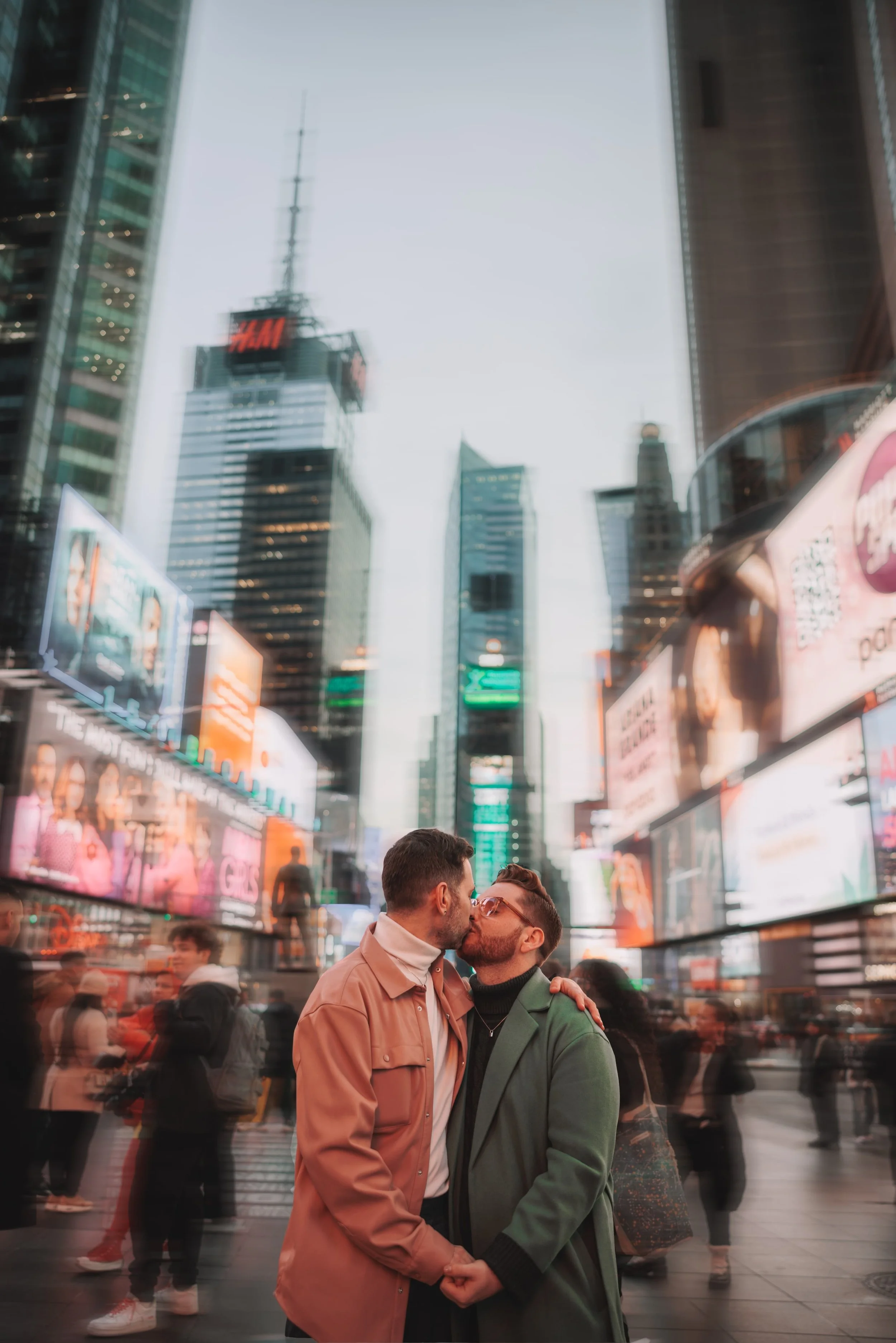 Two men kissing in Times Square, New York City, with illuminated billboards and tall skyscrapers in the background, surrounded by pedestrians.