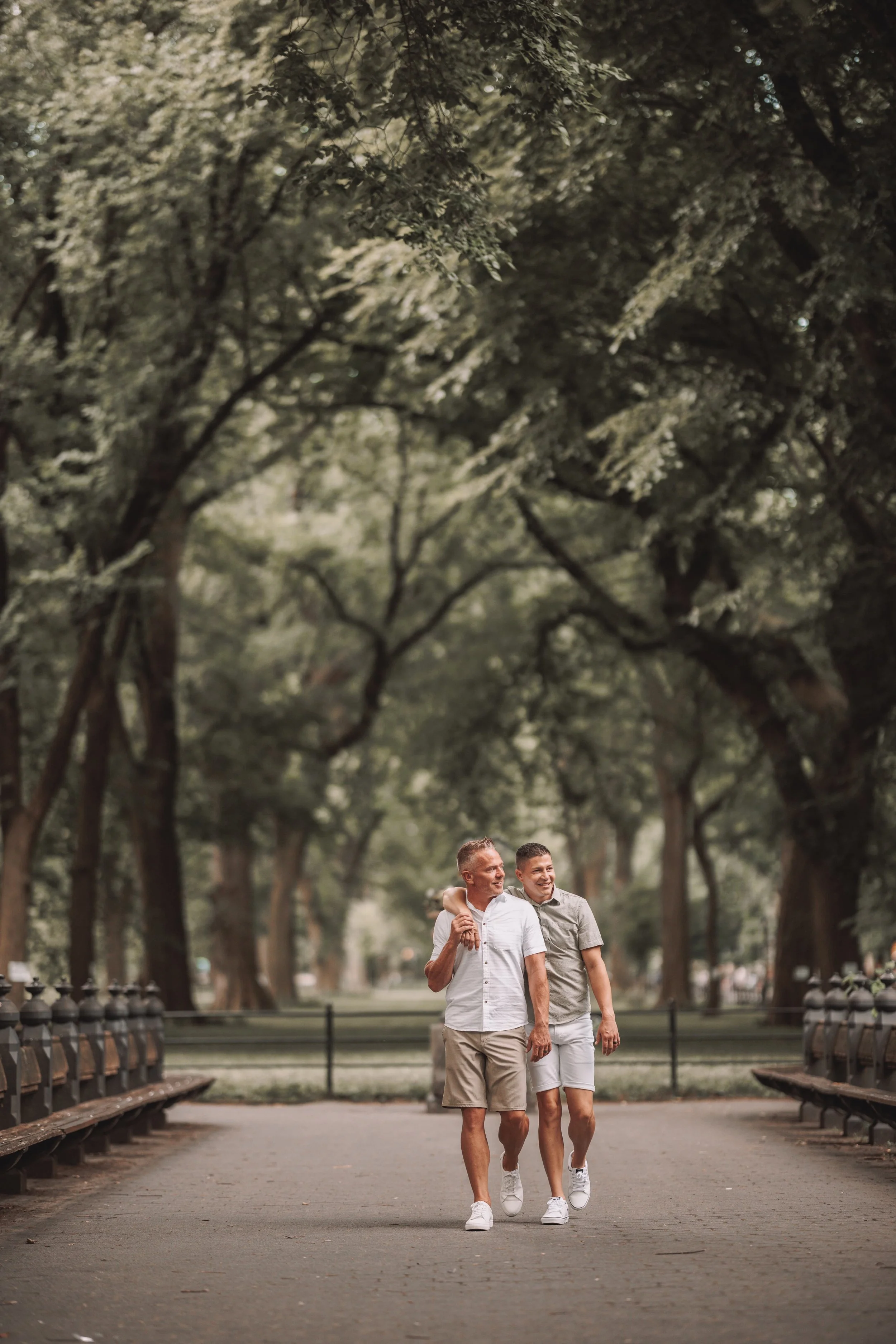 A man and a boy walking and smiling in a park with large trees and benches on both sides