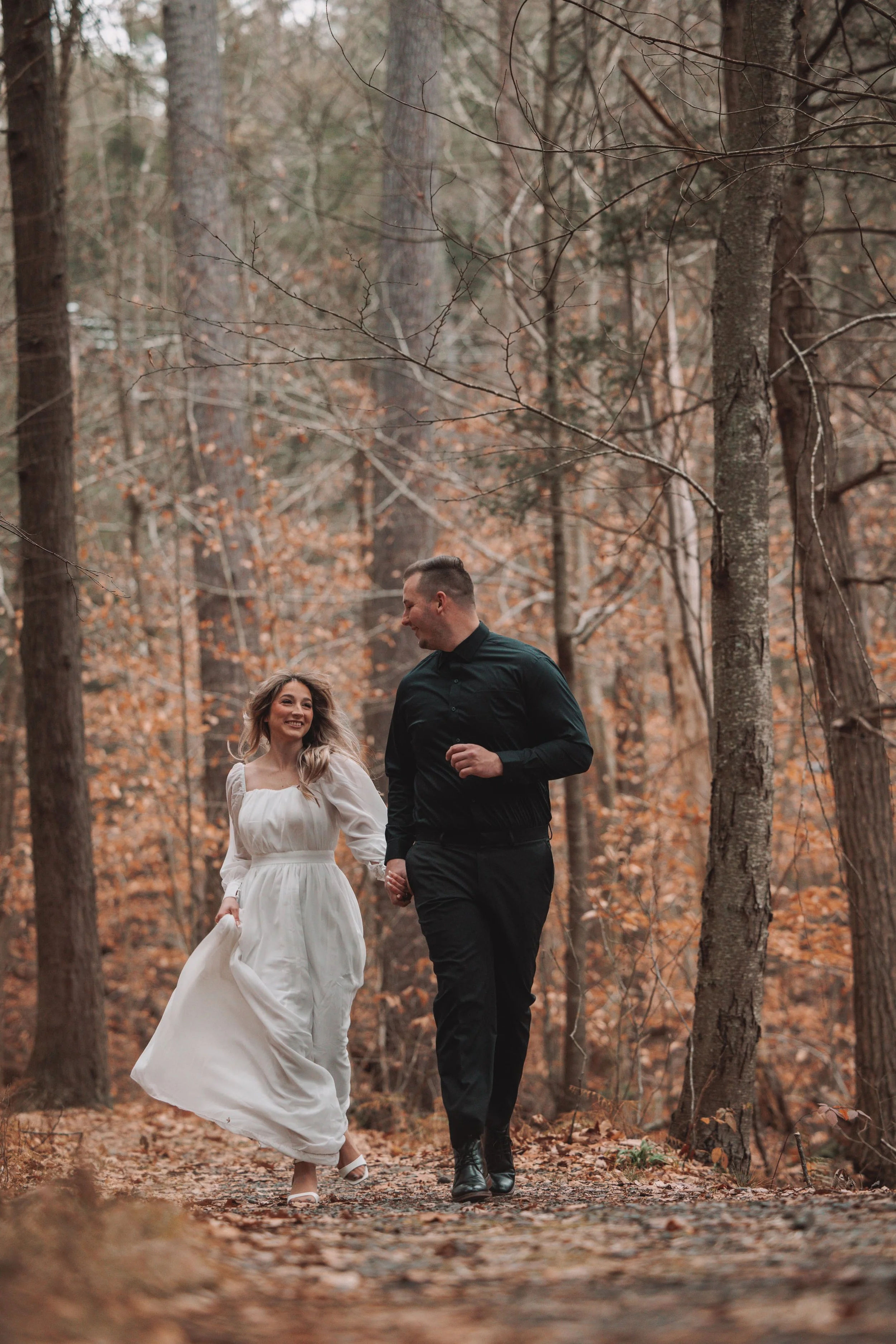 A couple, woman in a white dress and man in black, walking hand-in-hand through a wooded forest during autumn.