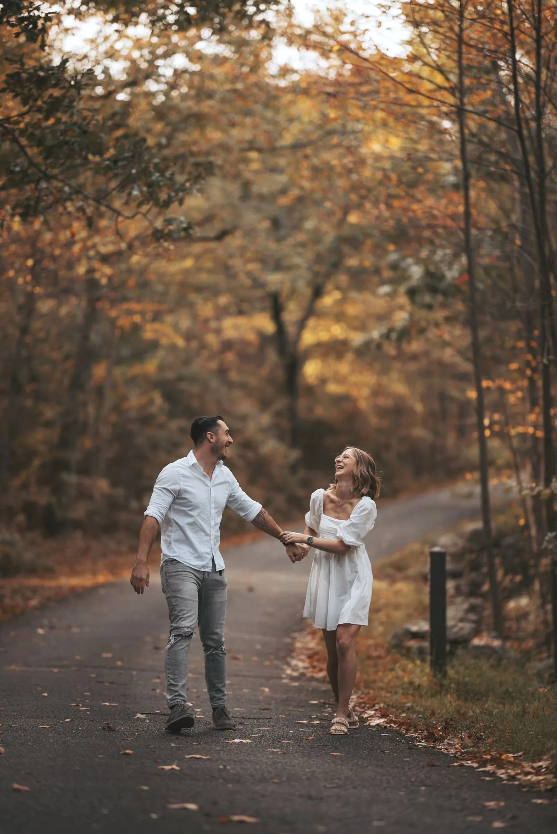 A smiling couple holding hands and walking along a scenic autumn trail surrounded by fall foliage, with trees and a winding path in the background.