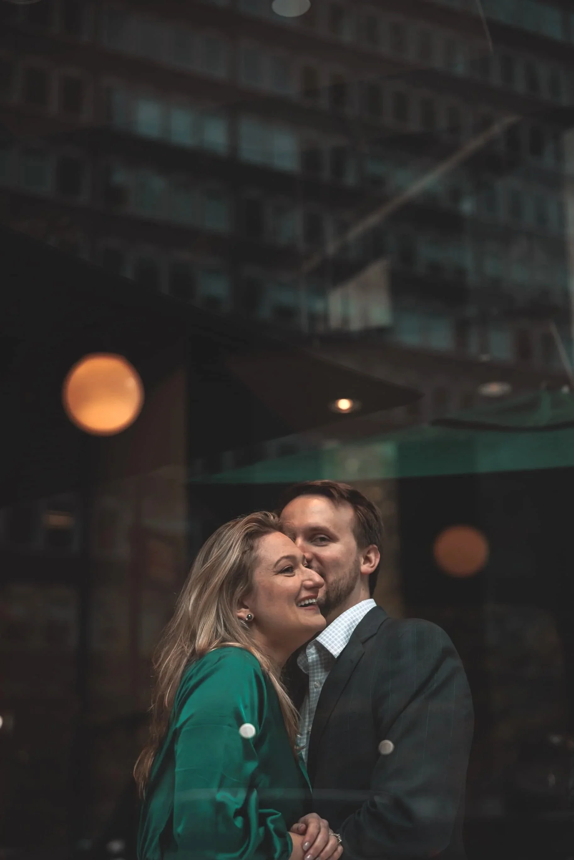 A couple shown through a window, smiling and embracing in an urban setting at night
