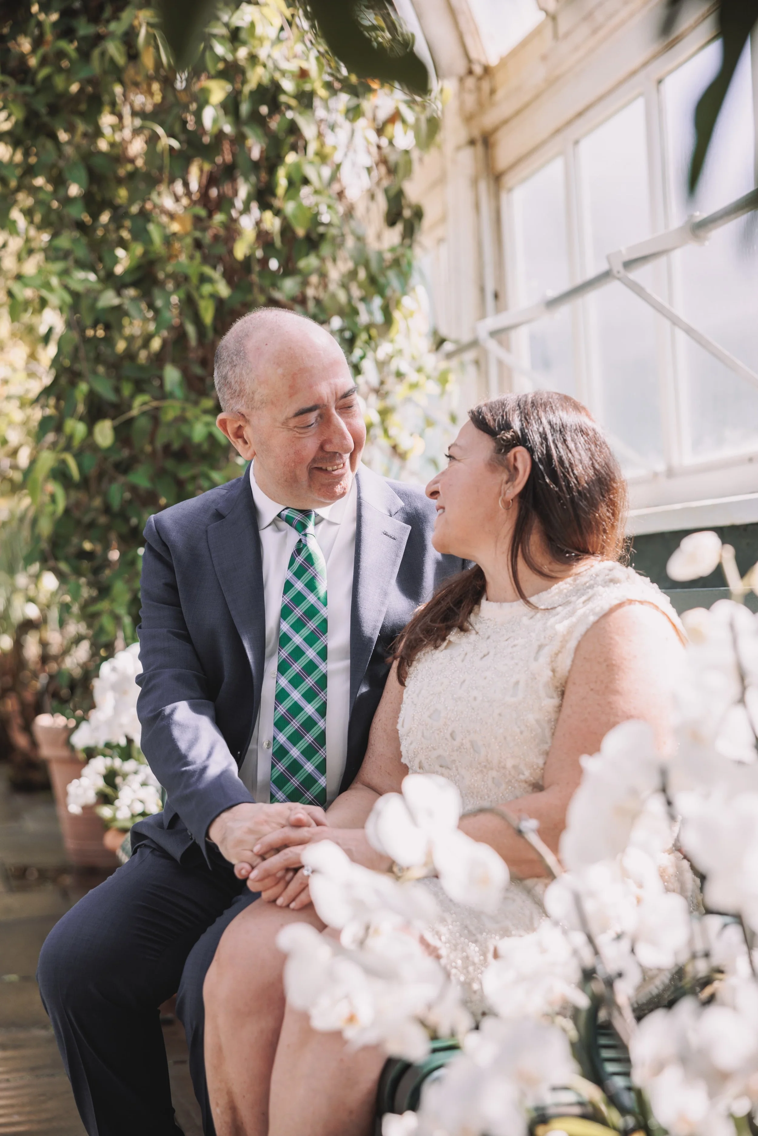 A couple holding hands and smiling at each other in a greenhouse with sunlight coming through the windows, surrounded by white flowers and plants.