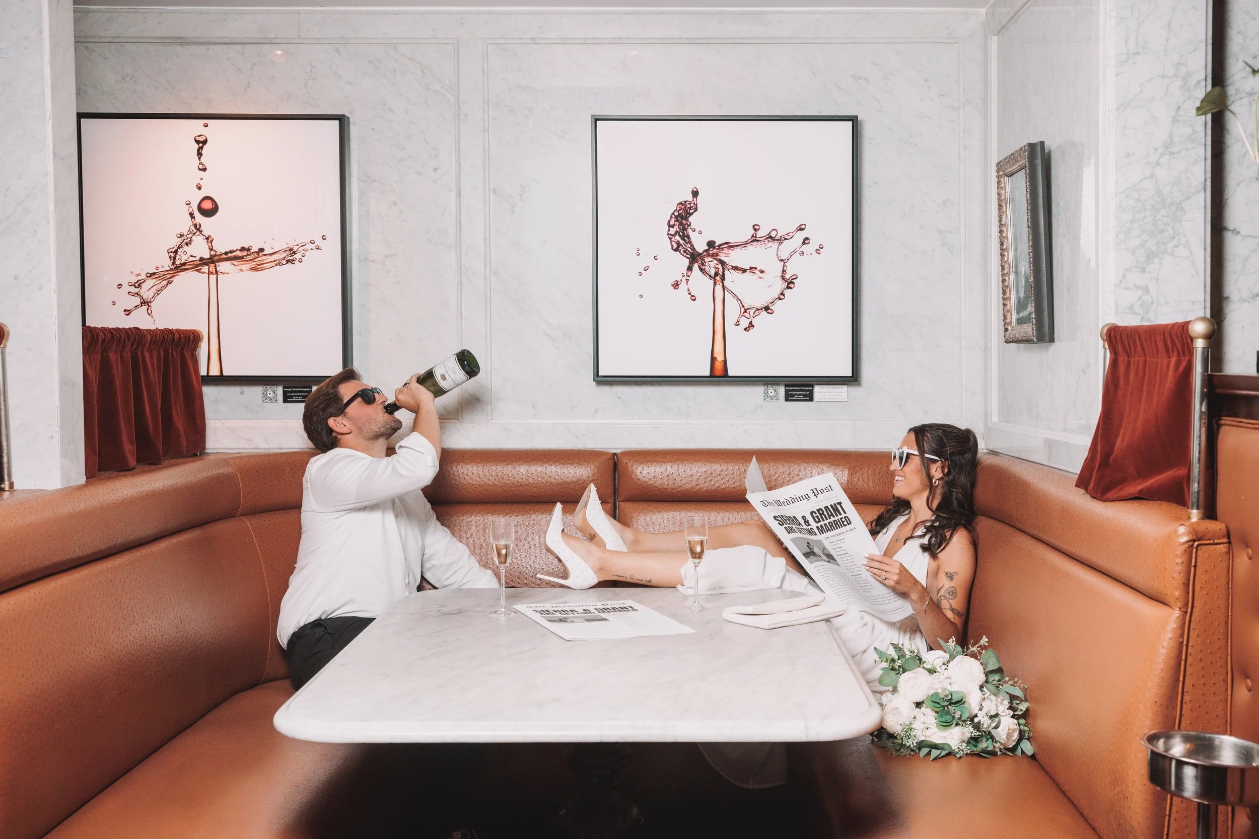 A couple sitting in a booth at a restaurant, with the woman holding a newspaper and the man drinking from a bottle, while the woman lounges with her feet on the table, and there are two glasses of champagne and a bouquet of flowers.