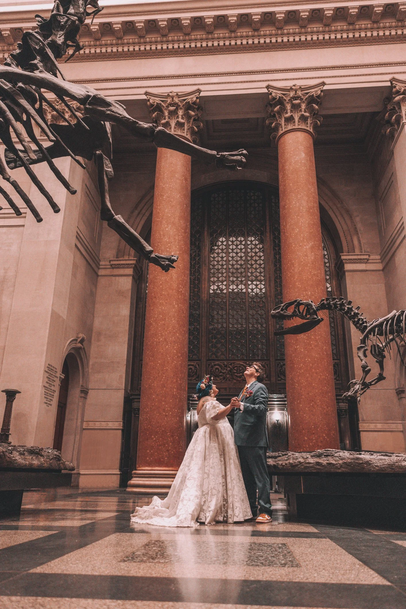 A bride and groom holding hands and gazing at each other inside a museum with large bronze dinosaur skeletons and grand columns.