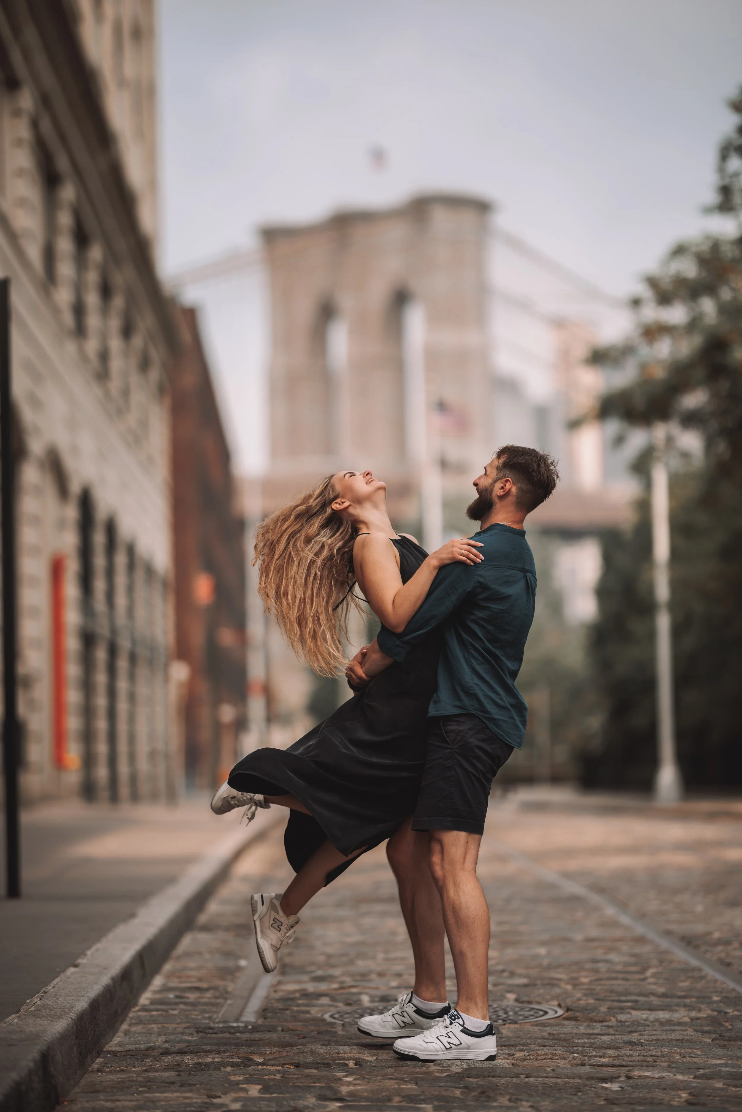A couple standing on a cobblestone street in New York City, with the Brooklyn Bridge in the background. The man is lifting the woman, and both are smiling and laughing.