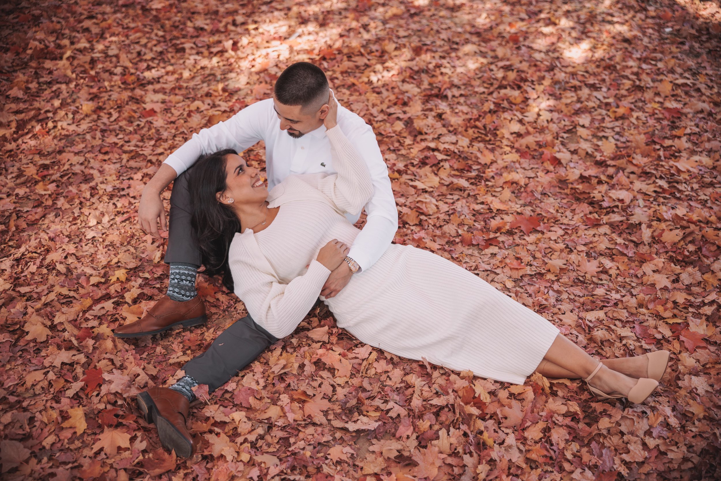 A couple lying on fallen autumn leaves, the woman wearing a cream dress and the man in a white shirt and dark pants, gazing at each other affectionately.