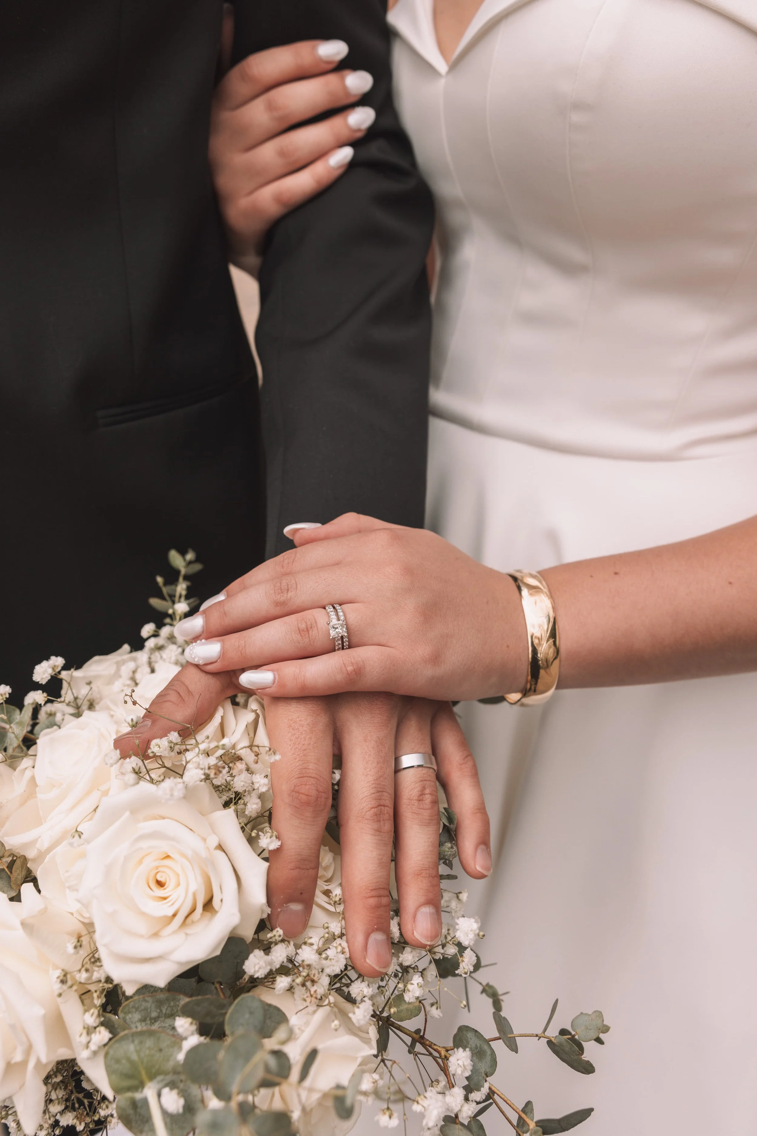 Close-up of a bride and groom's hands showing wedding rings, with a bouquet of white roses and greenery in the foreground.