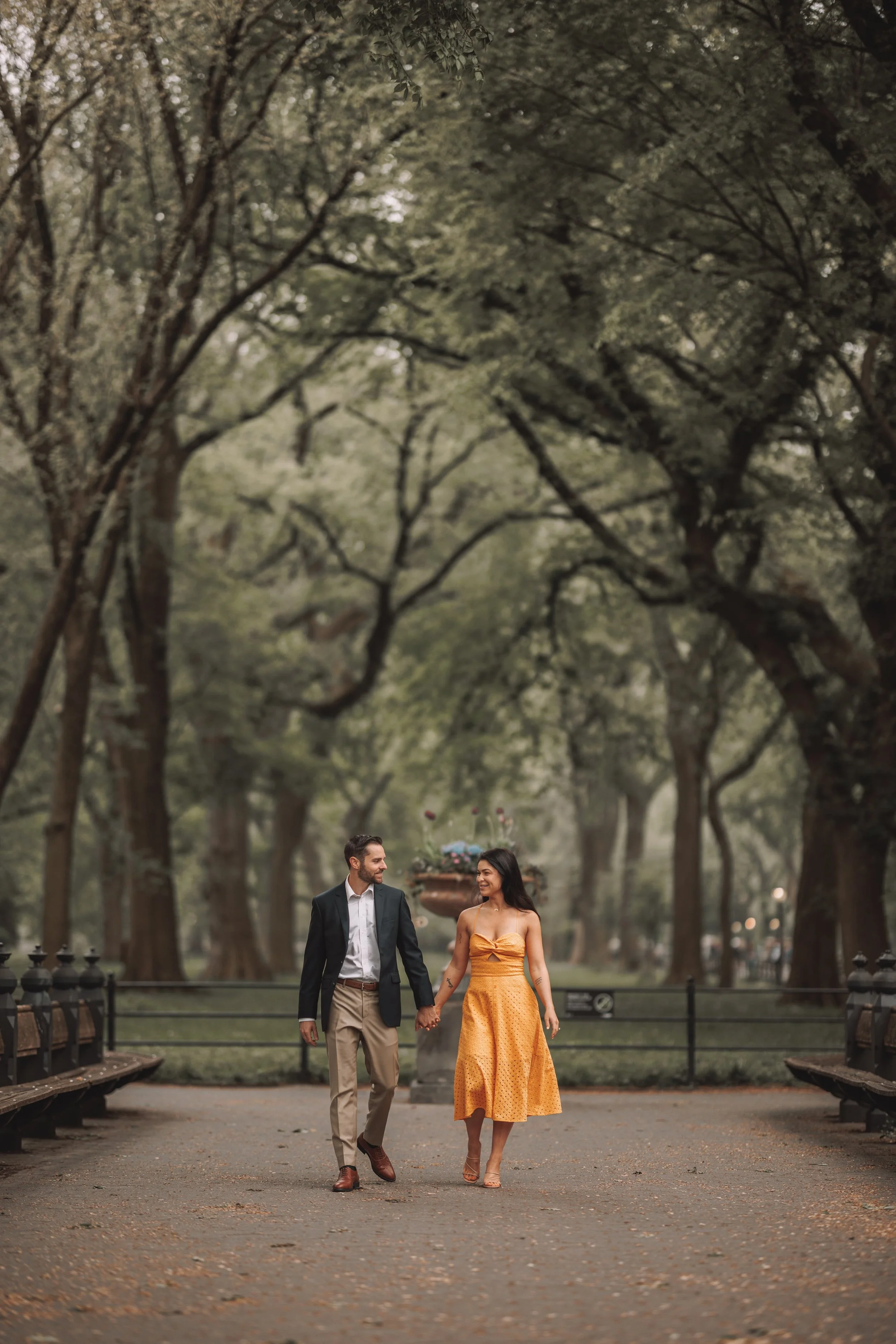 A man and woman are walking hand-in-hand down a park pathway surrounded by tall, leafy trees. The woman is wearing a yellow dress and heels, while the man is dressed in a suit jacket, shirt, and pants. They are smiling at each other as they walk.