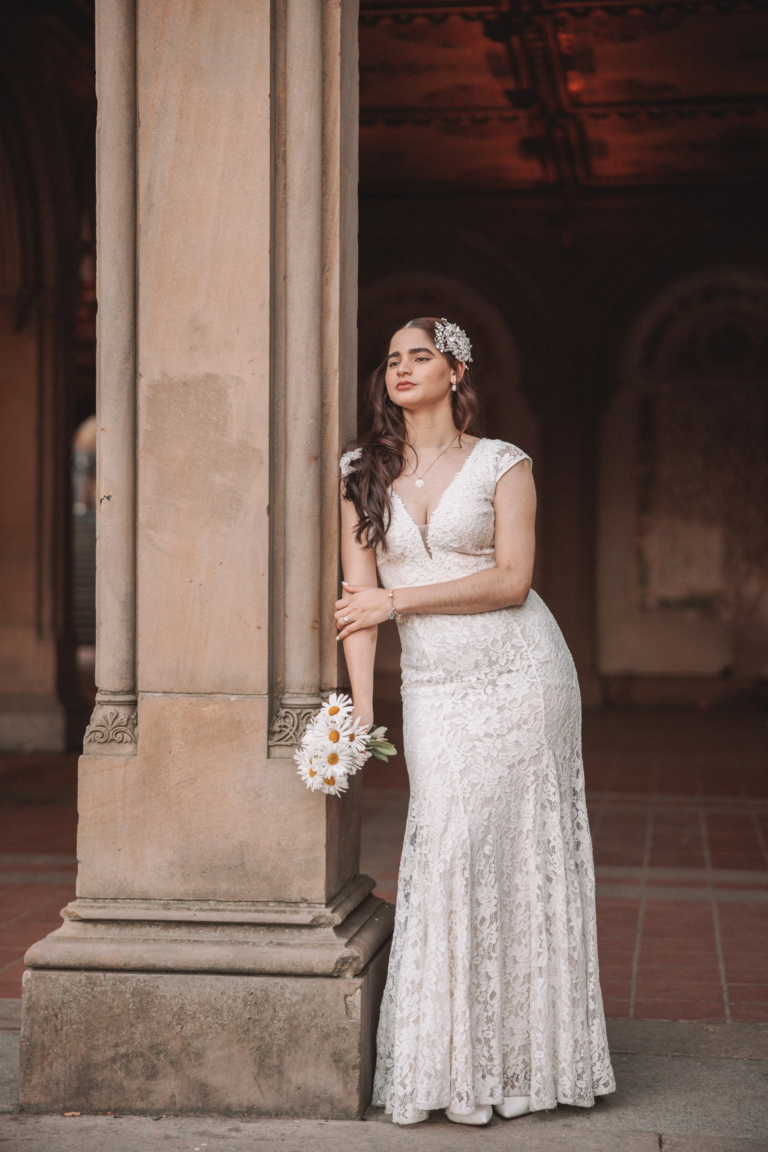 A woman in a white lace wedding dress standing beside a stone pillar, holding a bouquet of white daisies, with her eyes closed and a serene expression.