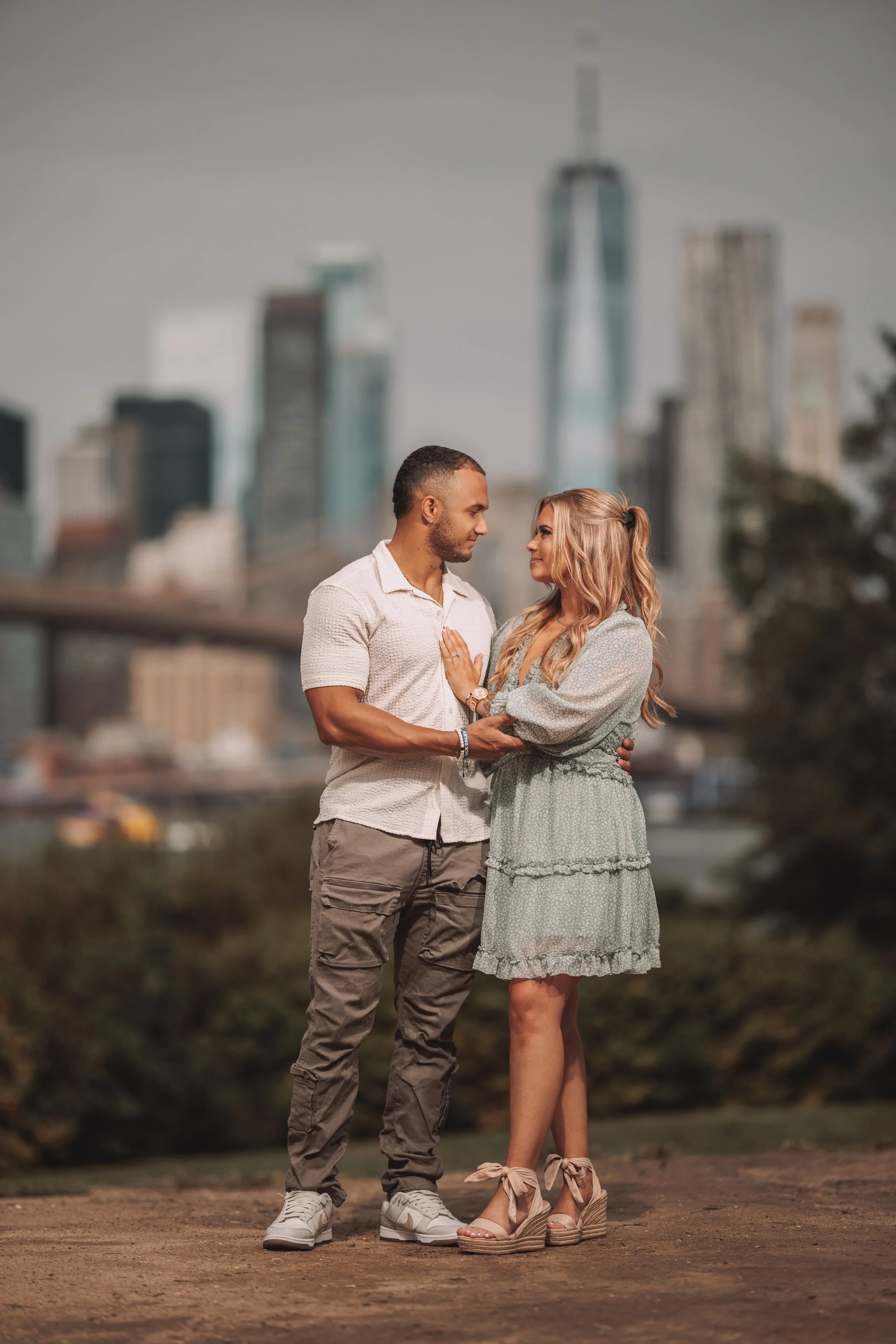 A young couple standing close together outdoors in front of a city skyline, with one person holding the other's arm and gazing into each other's eyes.
