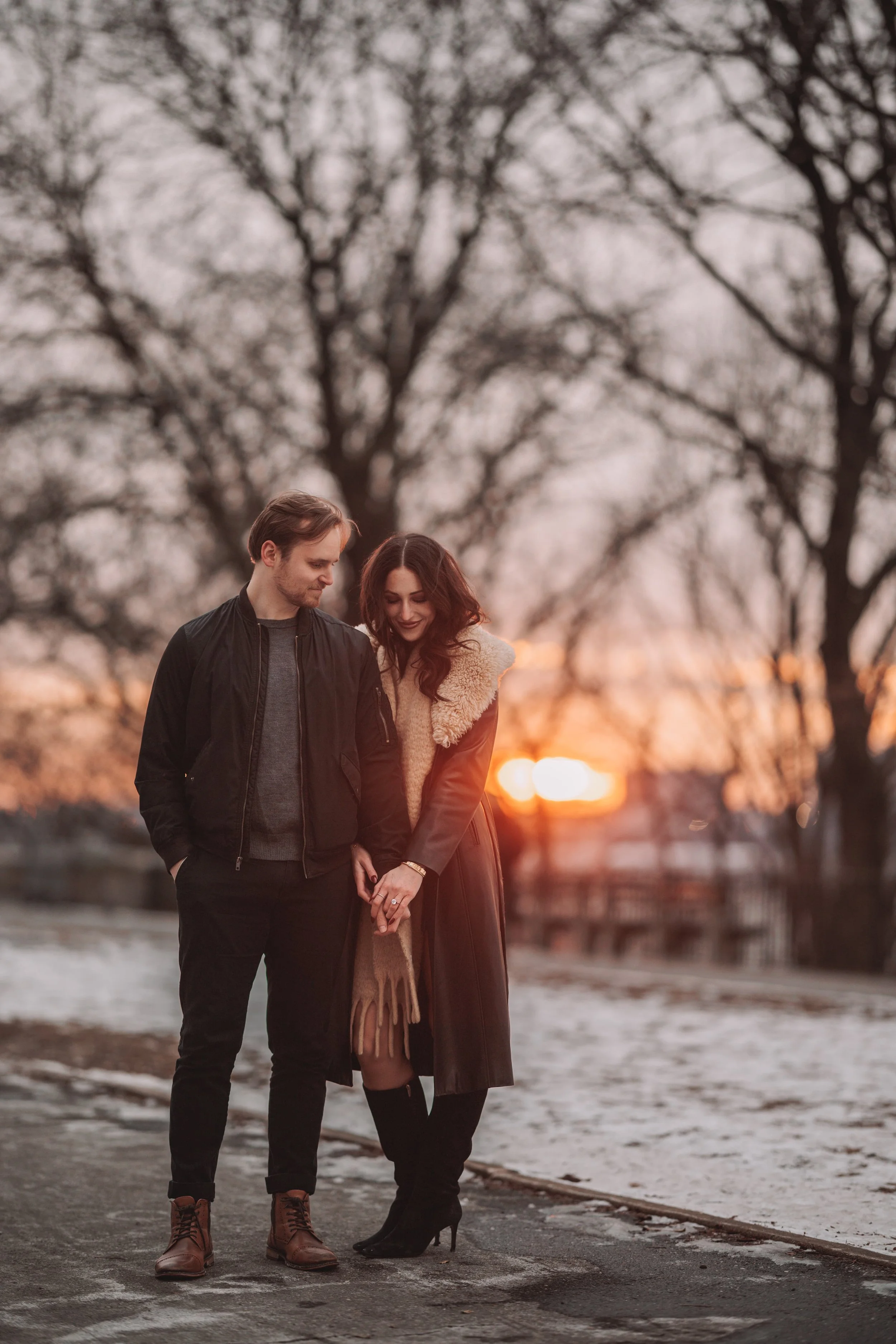 A couple holding hands and looking at each other outdoors at sunset with trees in the background.