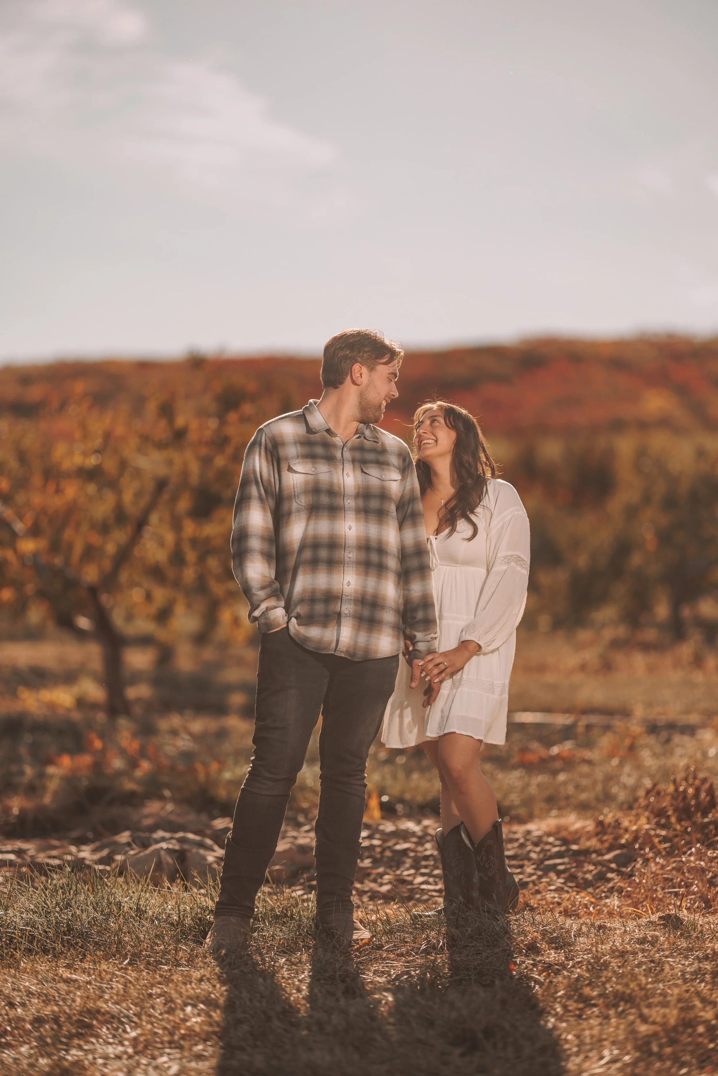A couple holding hands and smiling at each other outdoors during fall, with orange and yellow trees in the background and a cloudy sky.