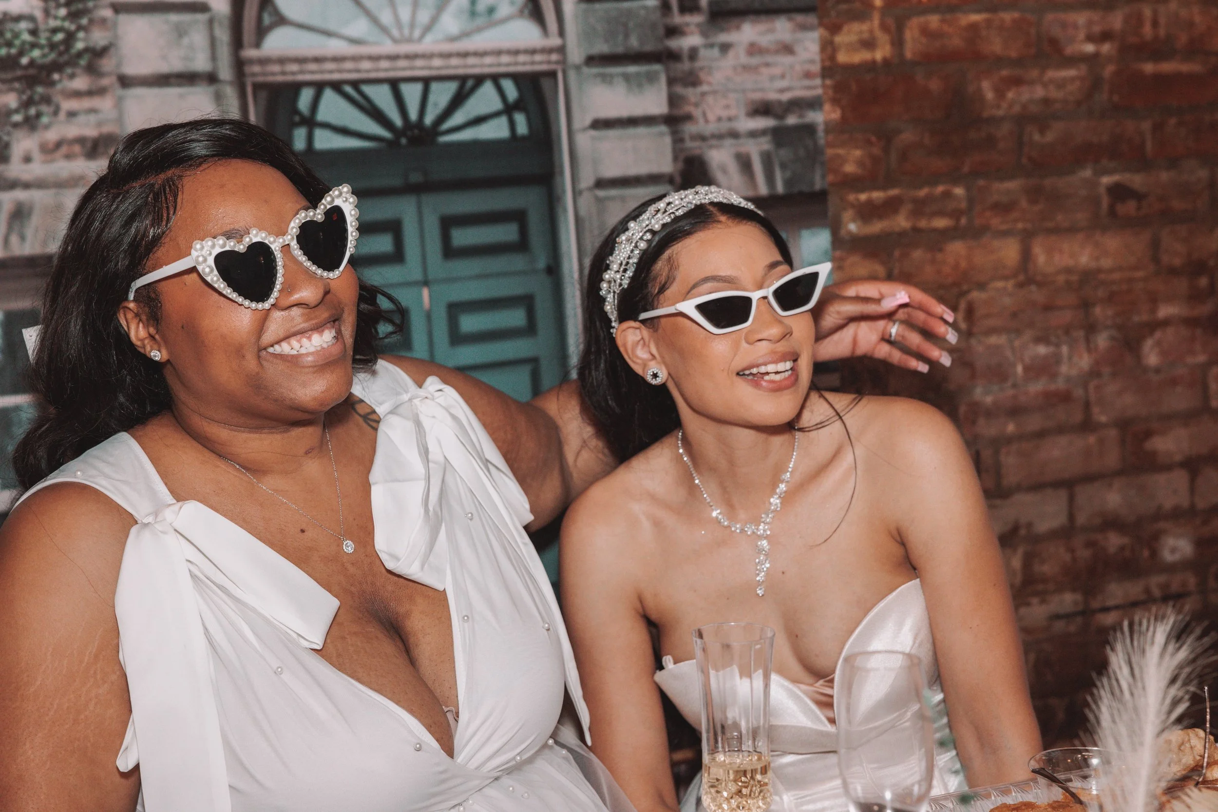 Two women at a celebration, wearing white dresses and sunglasses, smiling, with a brick wall and a decorative background behind them.