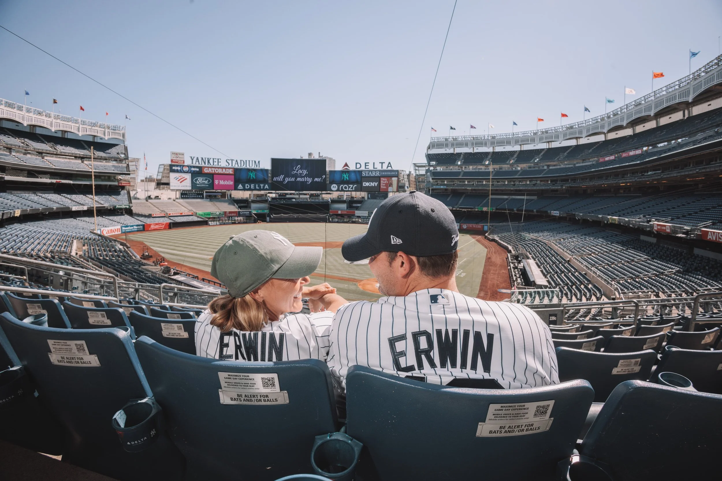 A couple sitting in Yankee Stadium seats, wearing New York Yankees jerseys and caps, overlooking the baseball field.