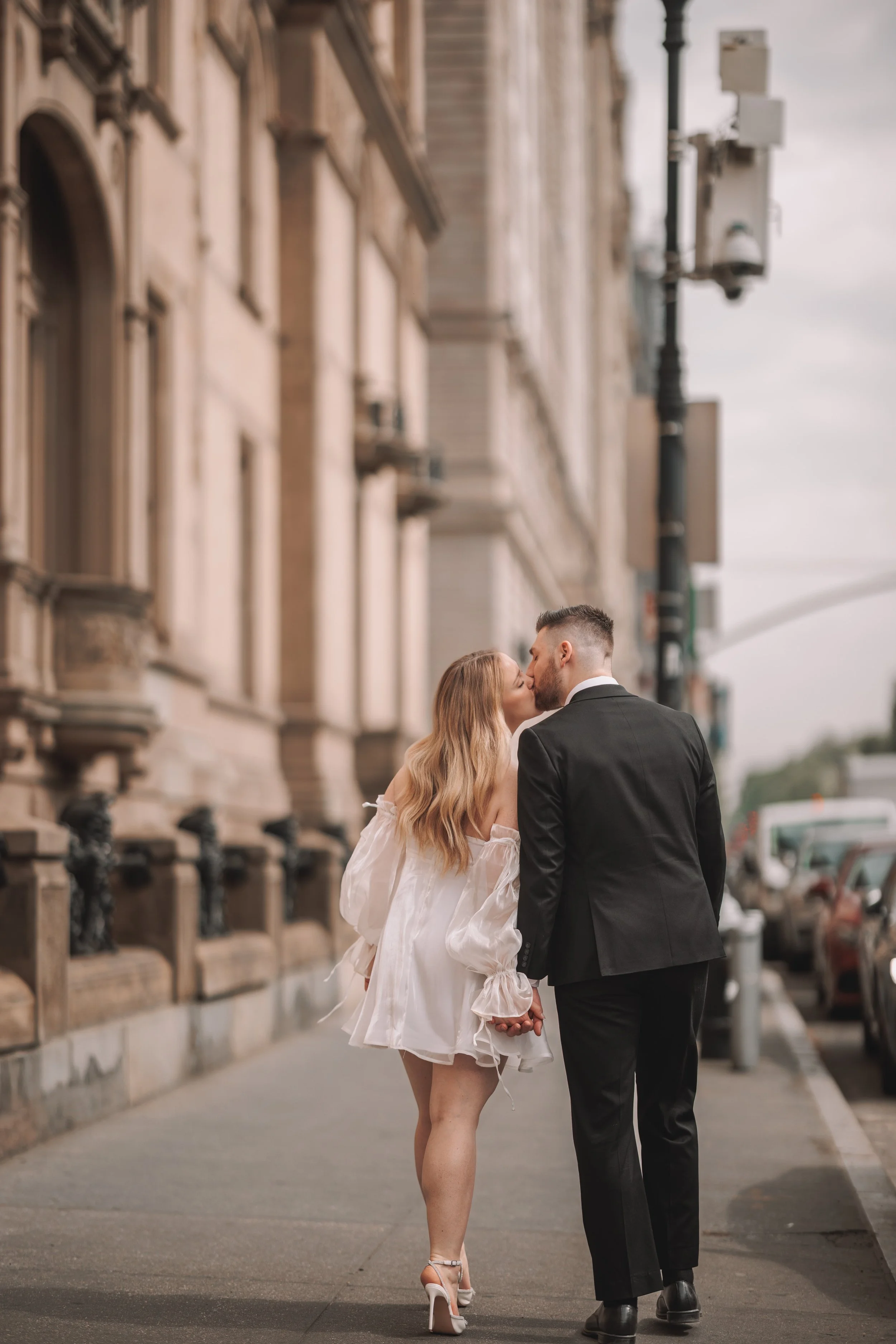 A couple wearing formal attire, with the woman in a white dress and high heels, and the man in a black suit, sharing a kiss and holding hands while walking on a city sidewalk.