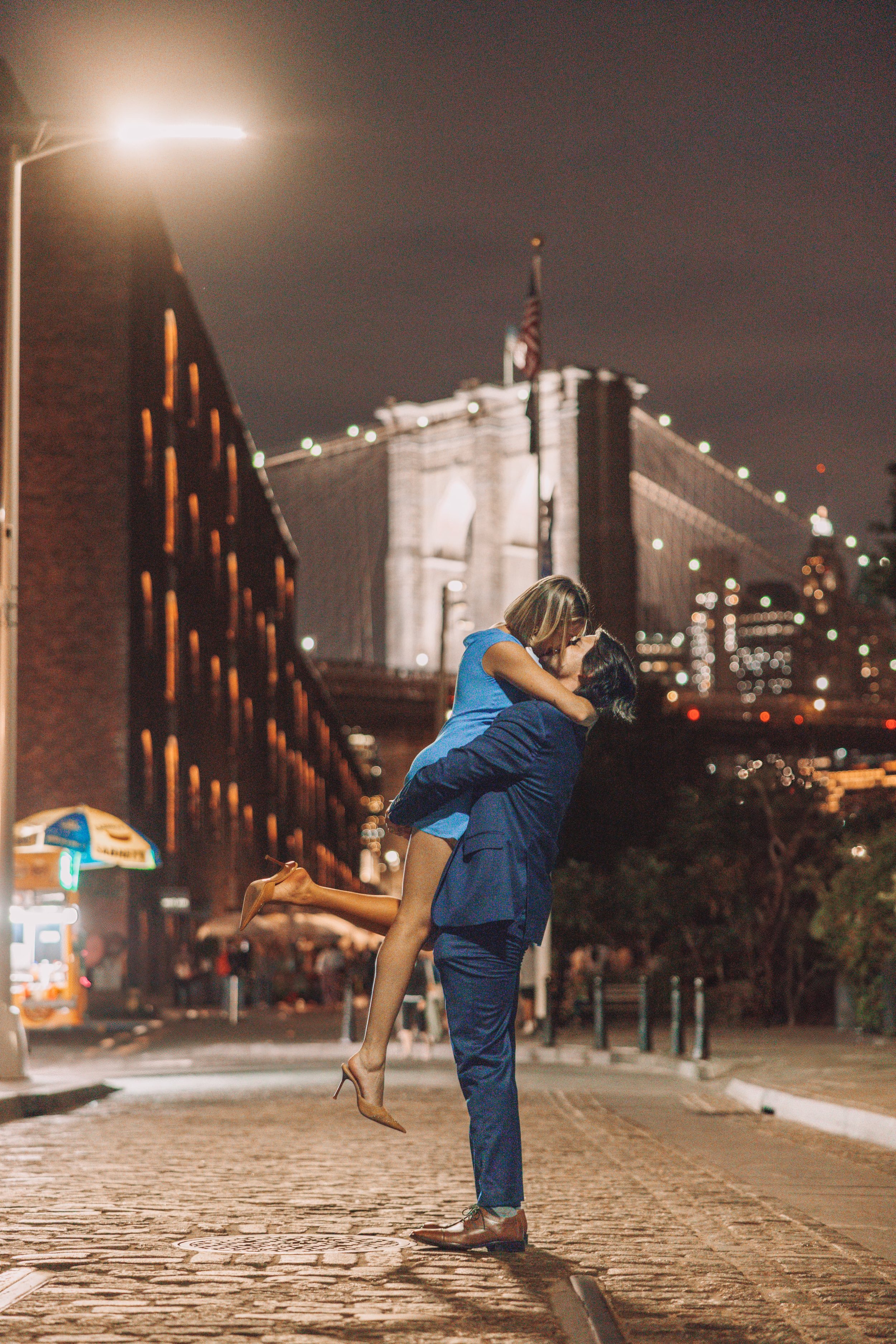 A man in a blue suit lifting a woman in a blue dress and heels in a romantic embrace on a city street at night, with the Brooklyn Bridge illuminated in the background.